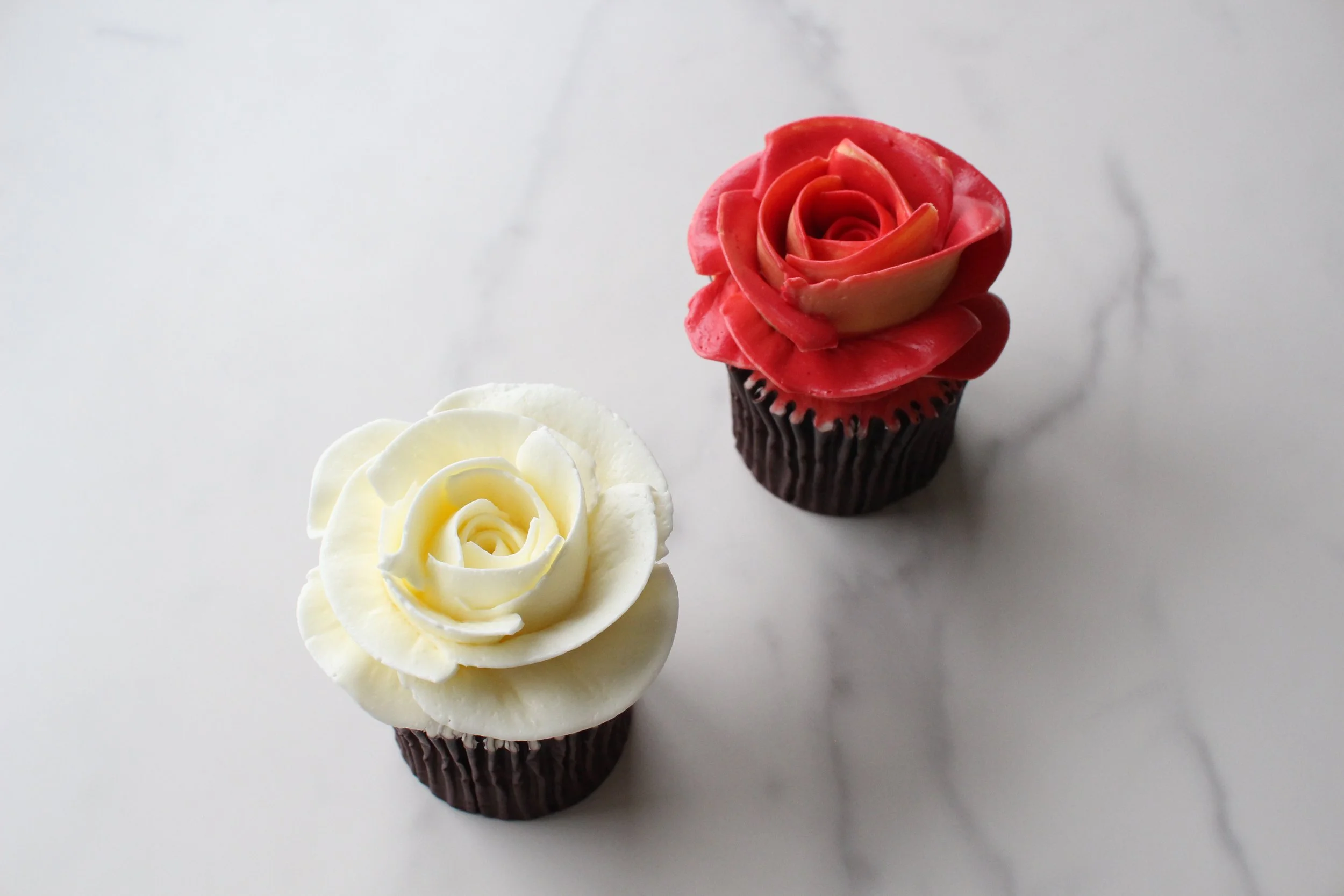 Red and white cupcakes decorated with rose-shaped frosting, placed on a white surface.