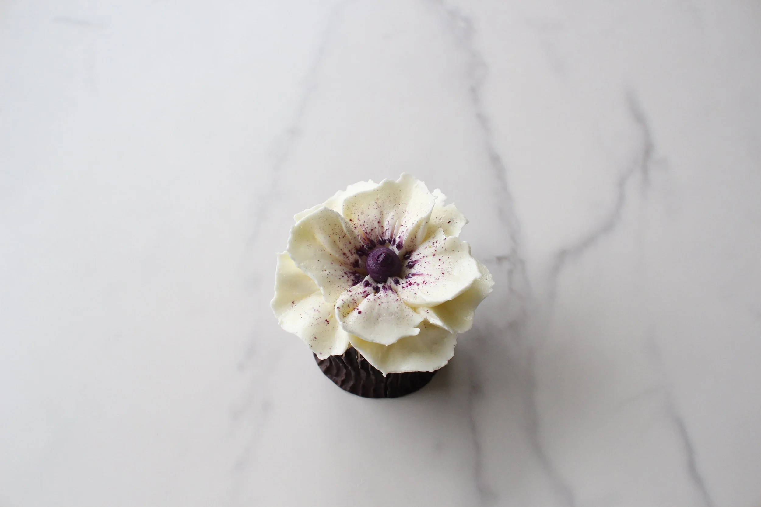 Cupcake with white frosting, purple edible flower, and dark chocolate base on a white surface.