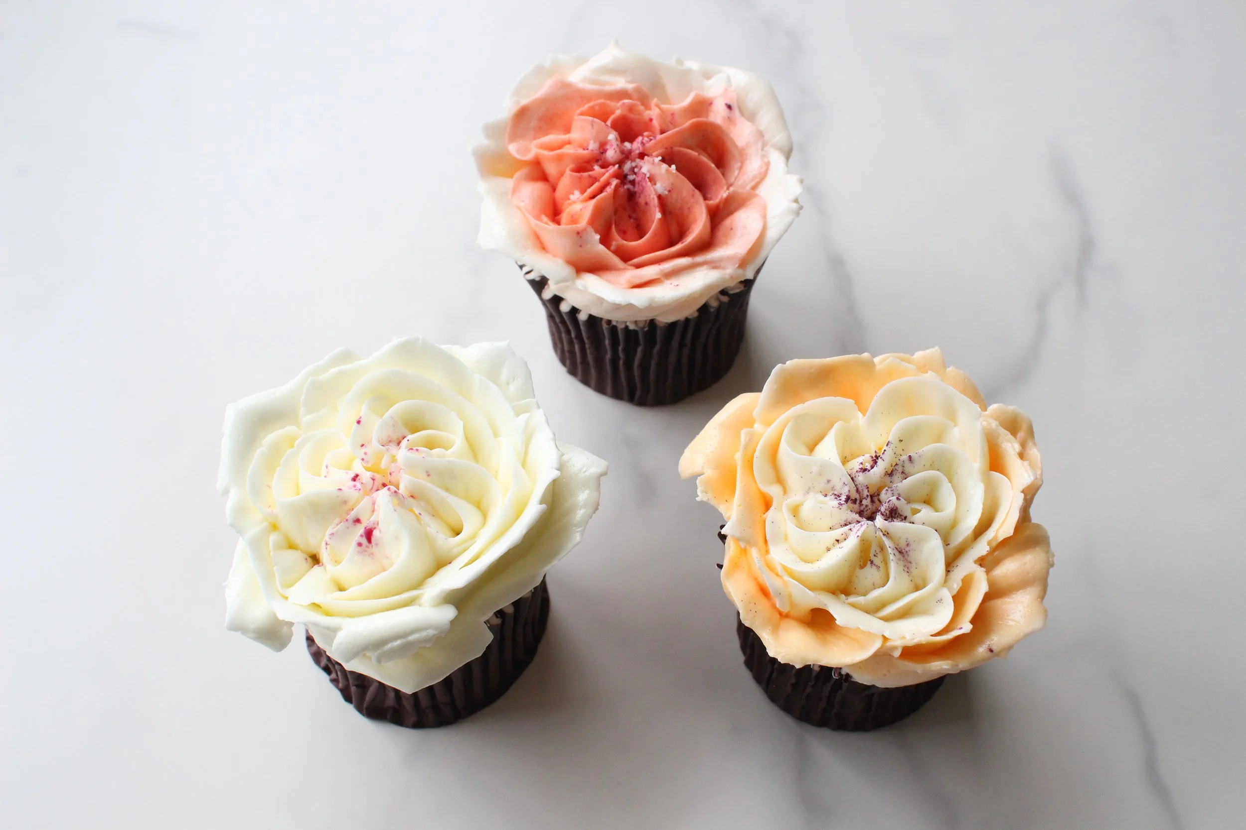 Four cupcakes with pastel-colored frosting designed to look like roses, on a white surface.