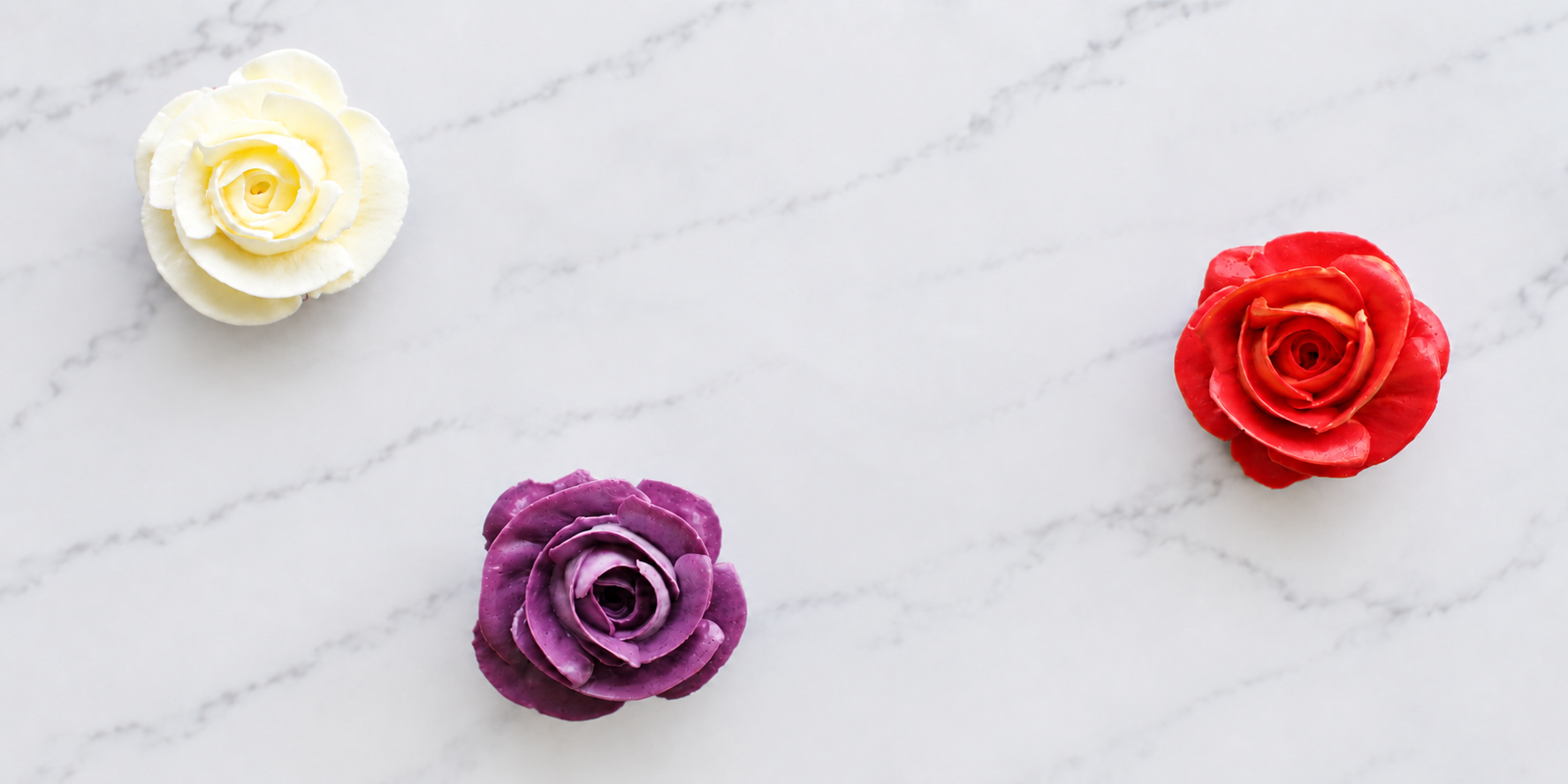 Three rose-shaped flowers in white, purple, and red color on a white marble surface.