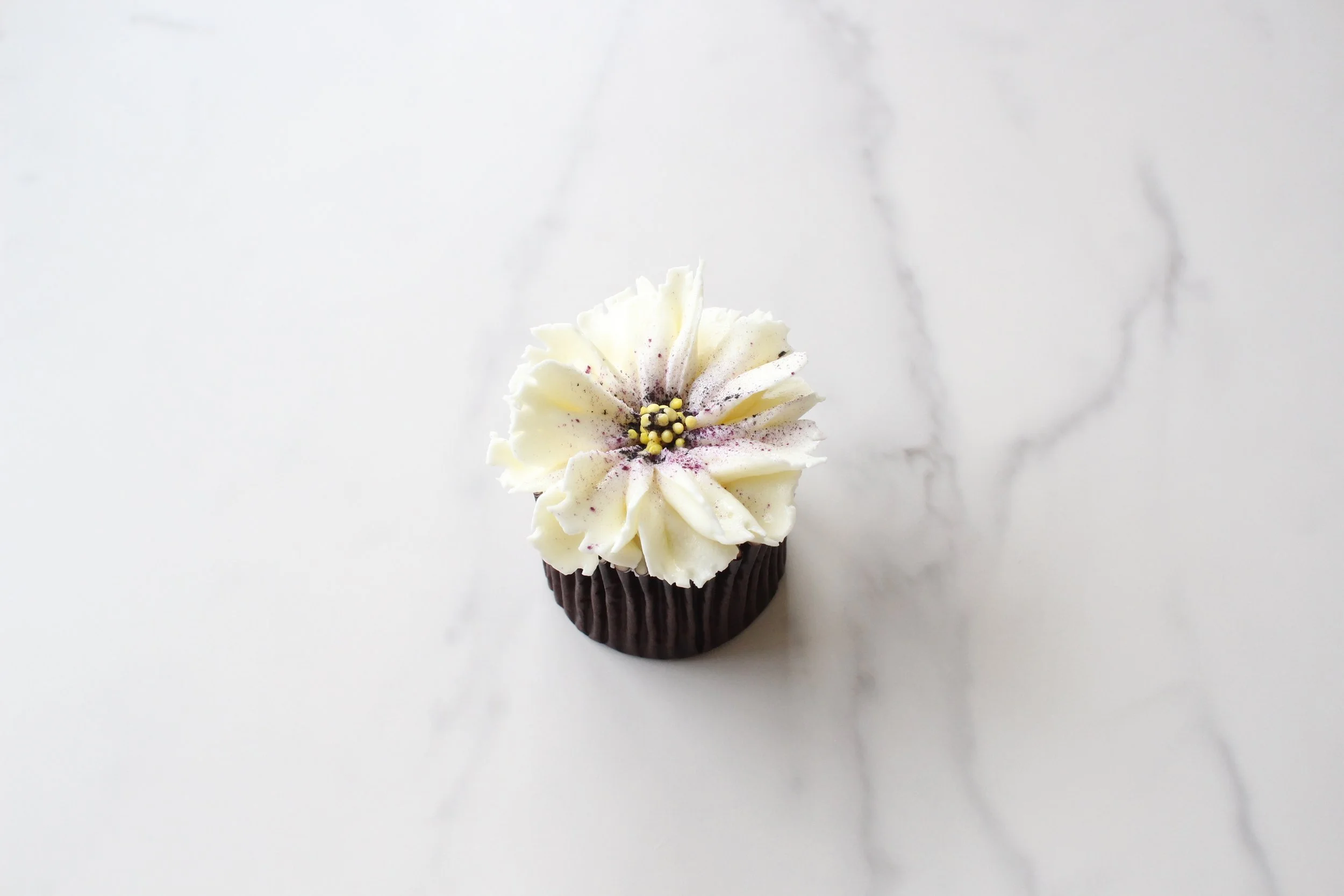 A cupcake with white and dark chocolate frosting decorated with purple sprinkles and yellow candy pearls, placed on a white marble surface.