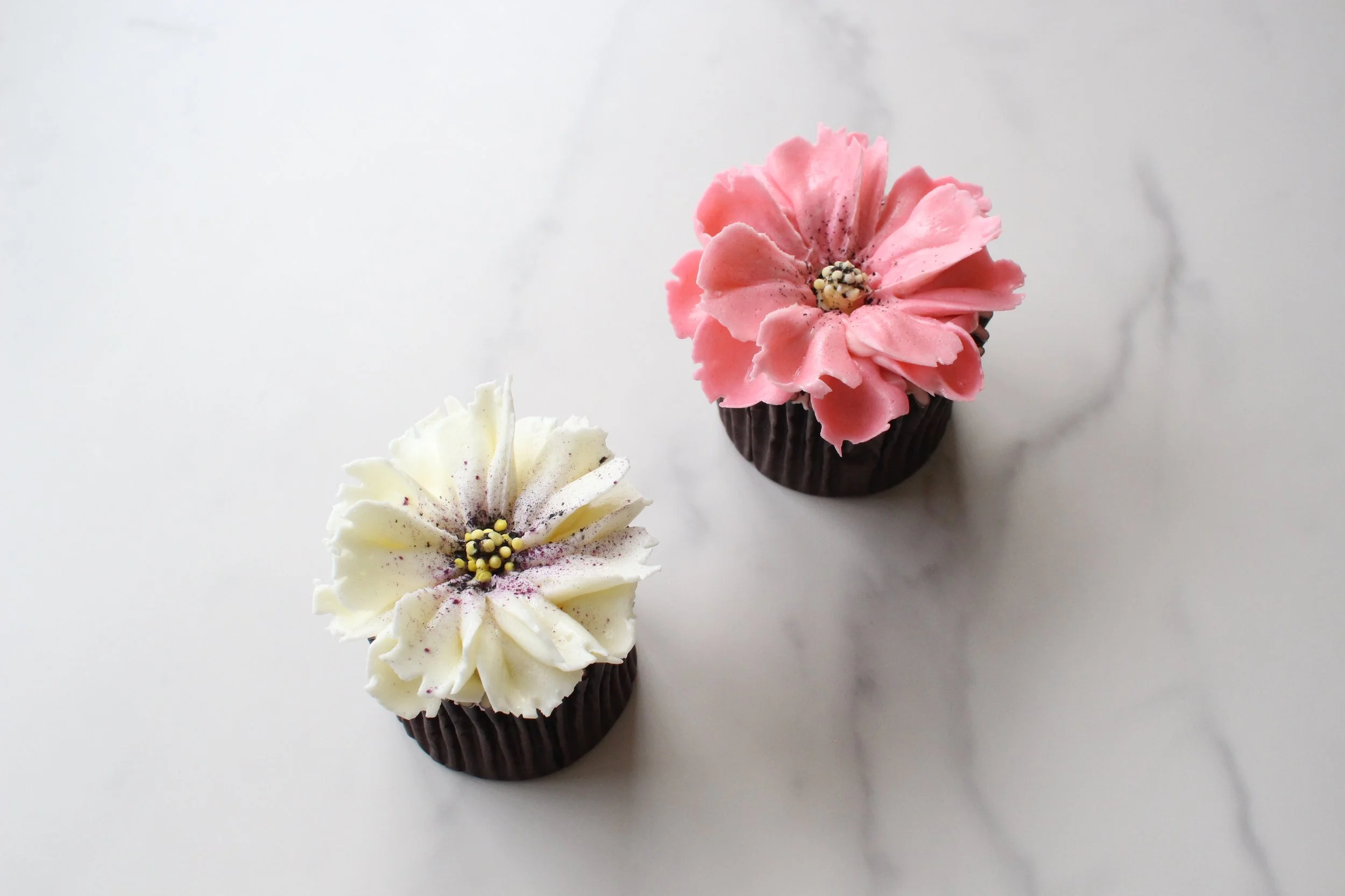 Two cupcakes with flower-shaped frosting, one pink and the other white, both topped with small edible decorations, placed on a white surface.