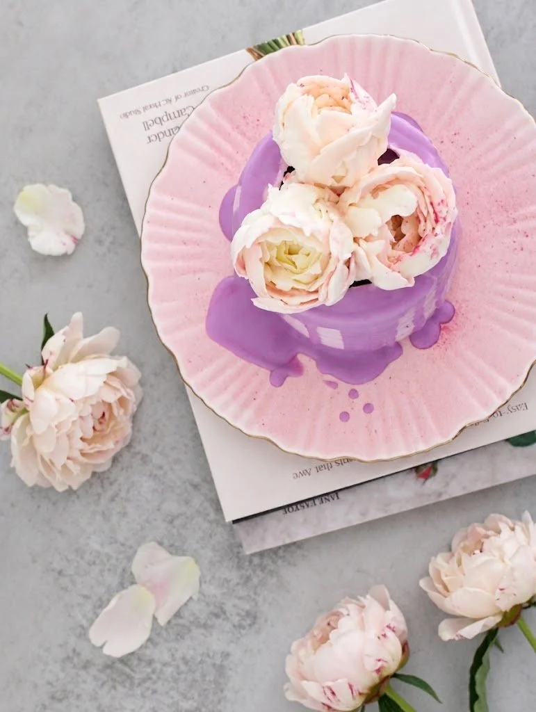 A pink ceramic plate with purple glaze accents holds several white and pink peony flowers. The plate is on top of a printed paper on a gray surface, with additional peony flowers and loose petals scattered around.