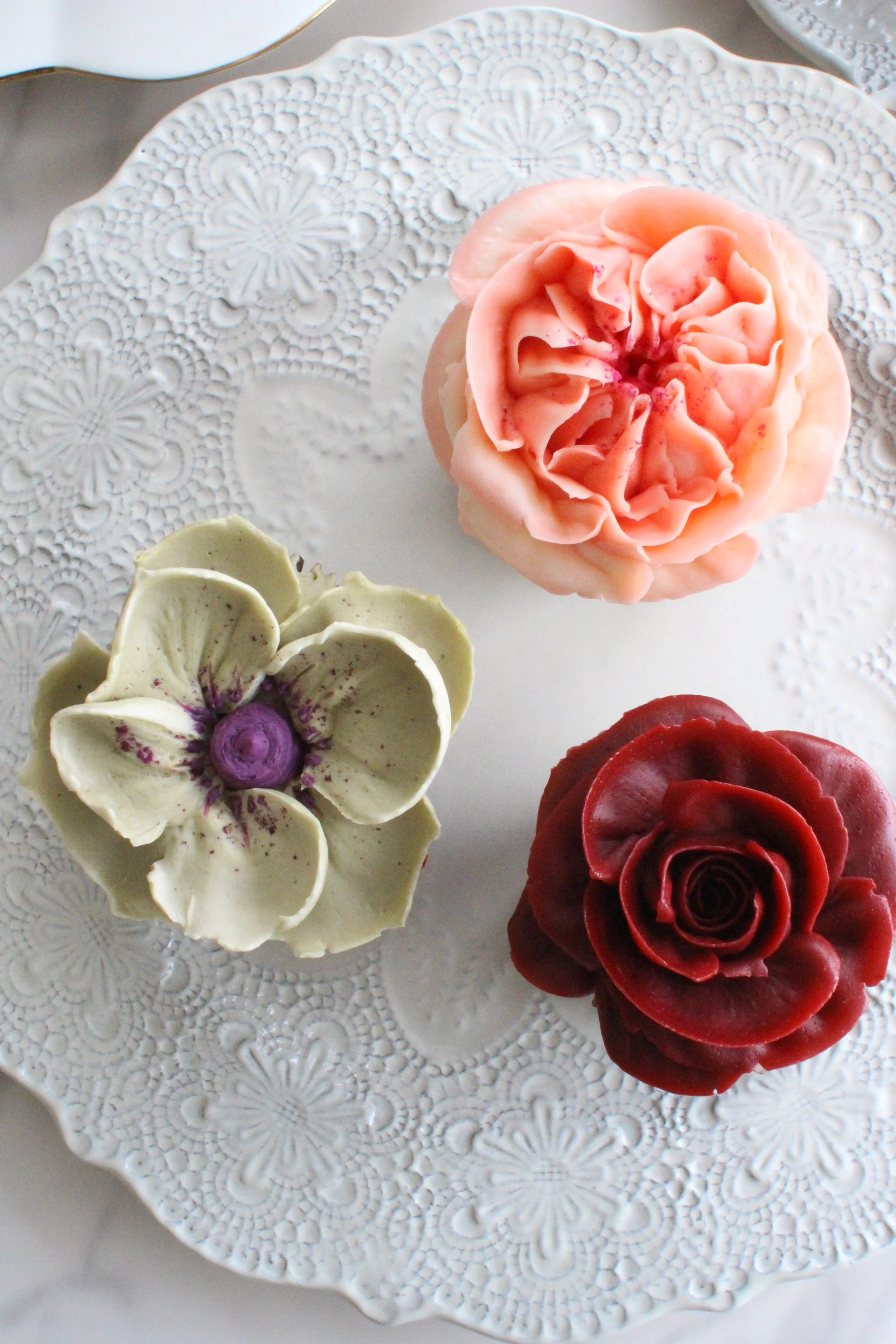 Assorted floral-shaped desserts on a decorative white plate, including a pink rose-shaped pastry, a white flower with purple accents, and a deep red rose-shaped pastry.