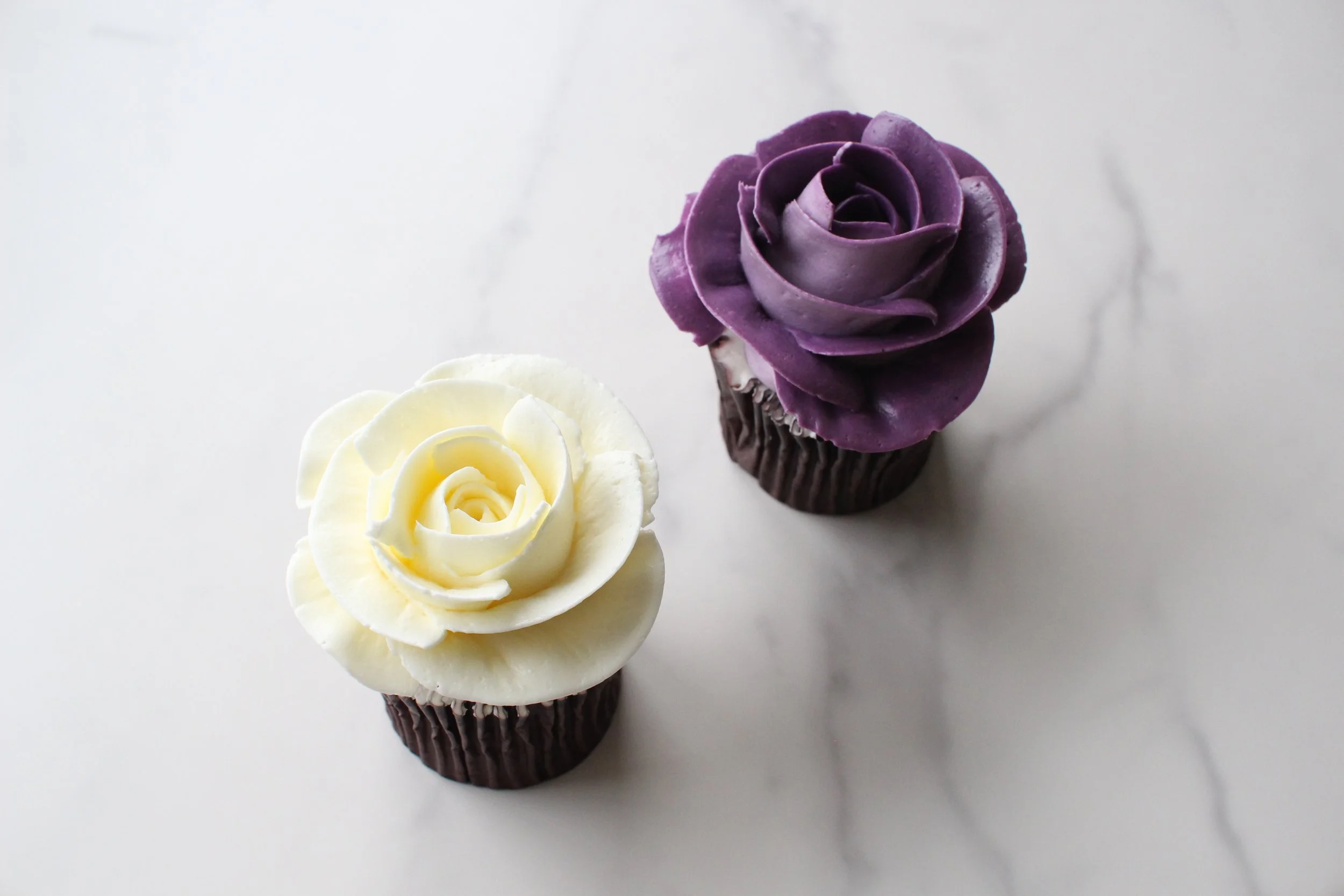 Two cupcakes decorated with rose-shaped frosting, one with white frosting and the other with purple frosting, placed on a white surface.