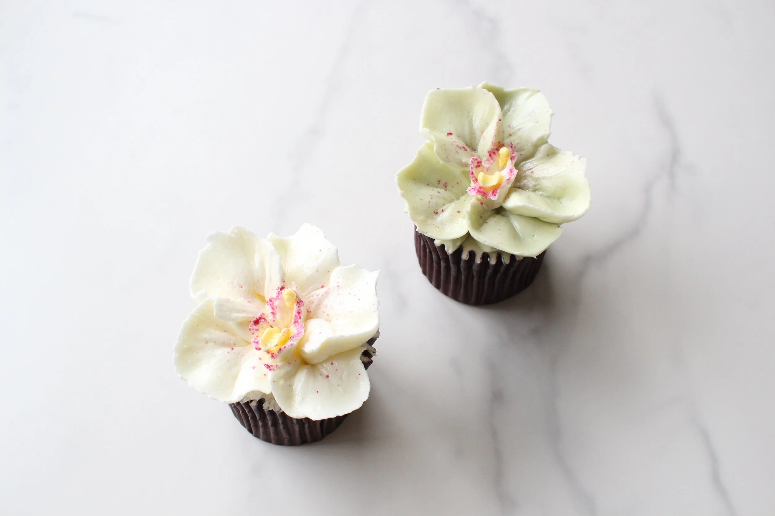 Two cupcakes with light green and white icing decorated to resemble orchid flowers, set on a white surface.
