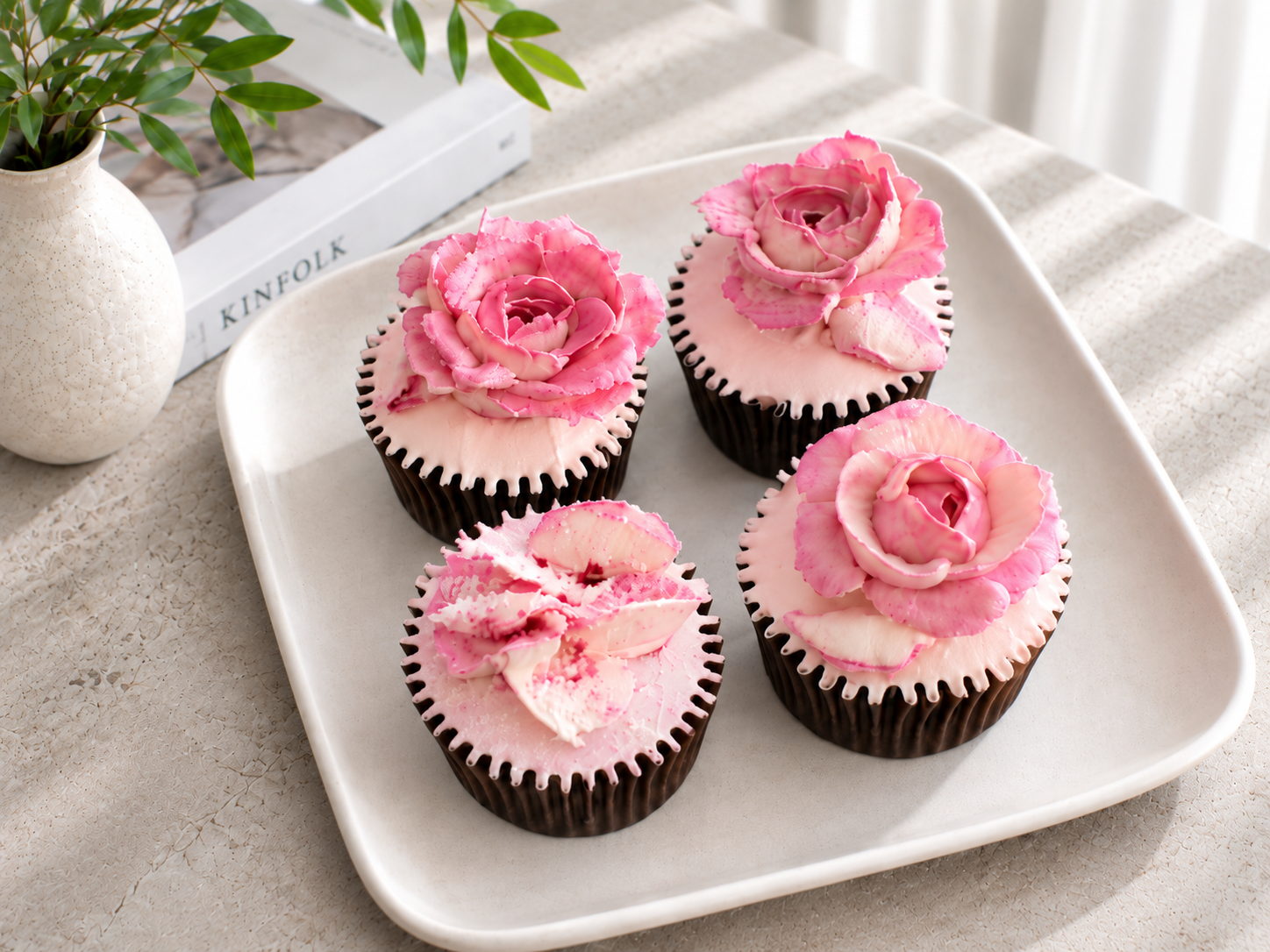 Four cupcakes with pink and white frosting roses on top, placed on a white rectangular plate.