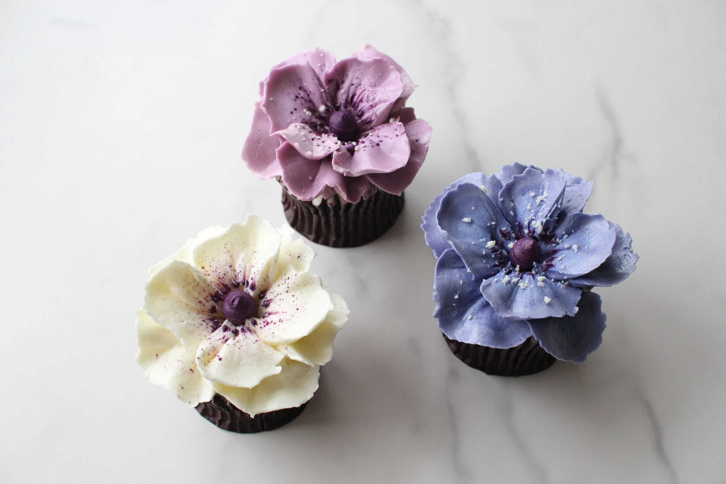 Decorative cupcakes with flower-shaped frosting in pastel colors of white, yellow, pink, blue, and purple, each topped with a small round confection resembling a flower center, arranged on a white surface.