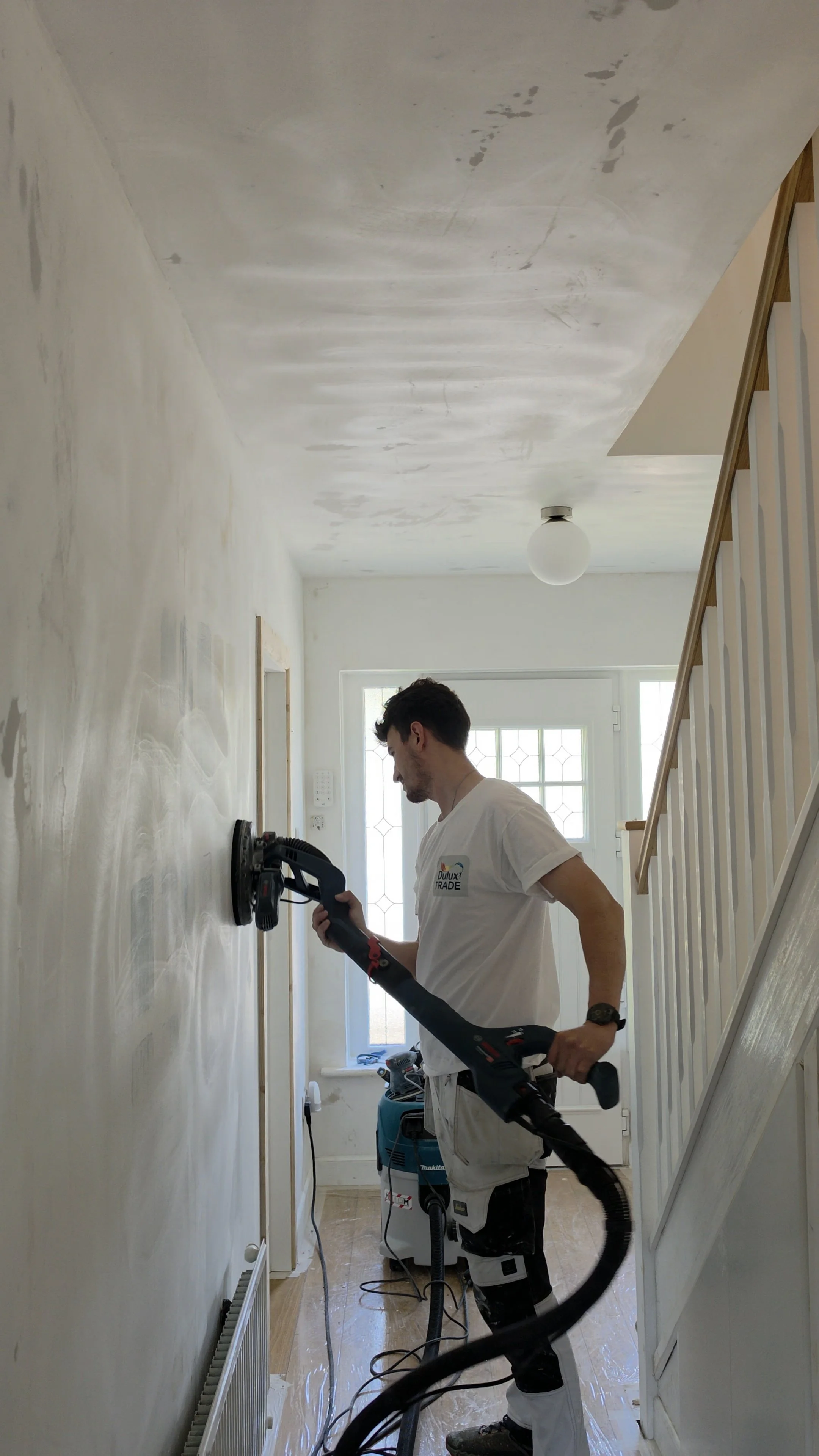A man using a power sander on a wall inside a house, with a staircase on his right and a door with a window behind him.