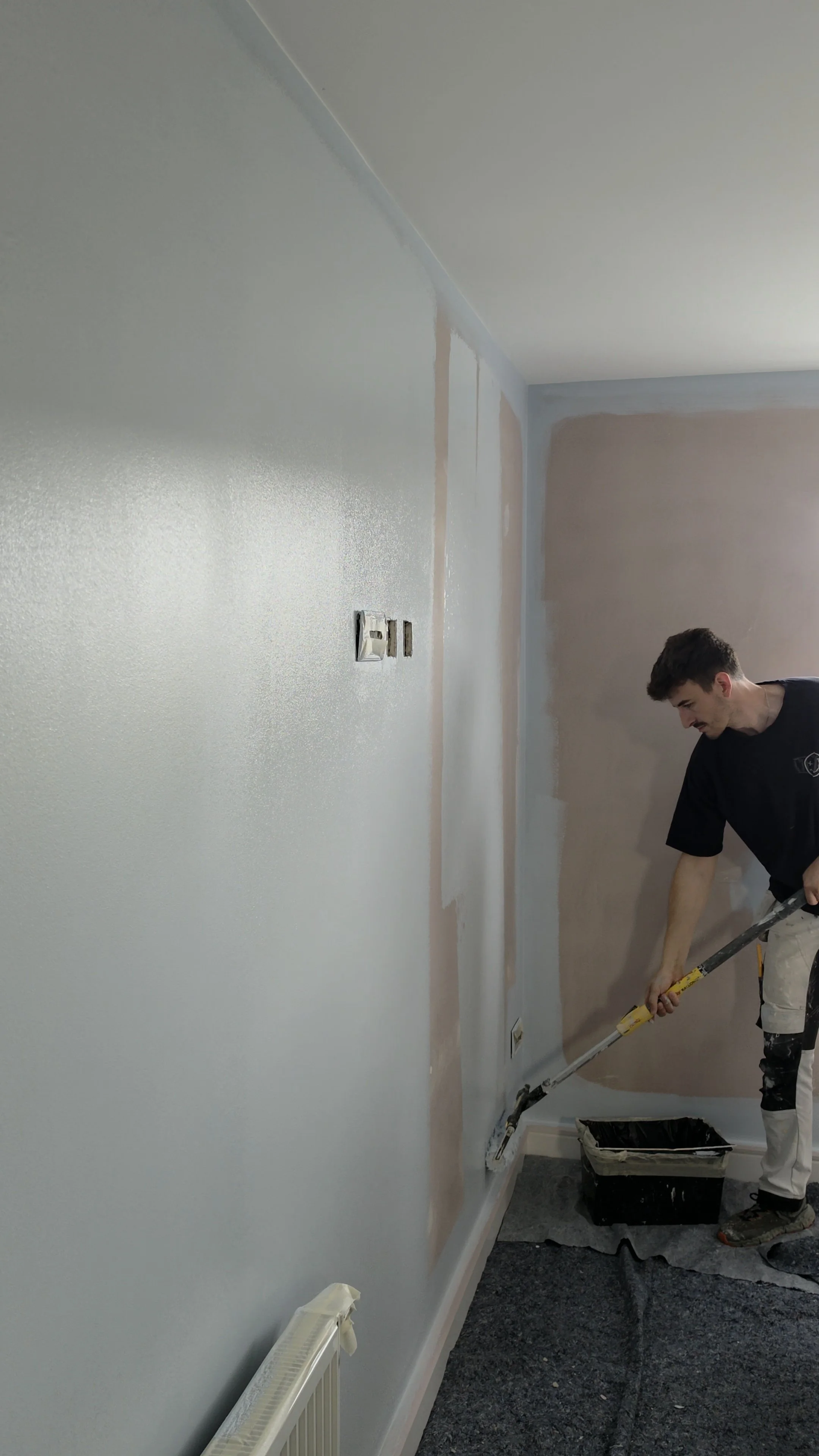 A person painting a wall with a roller, with sections of primer and paint visible, in a room under renovation.
