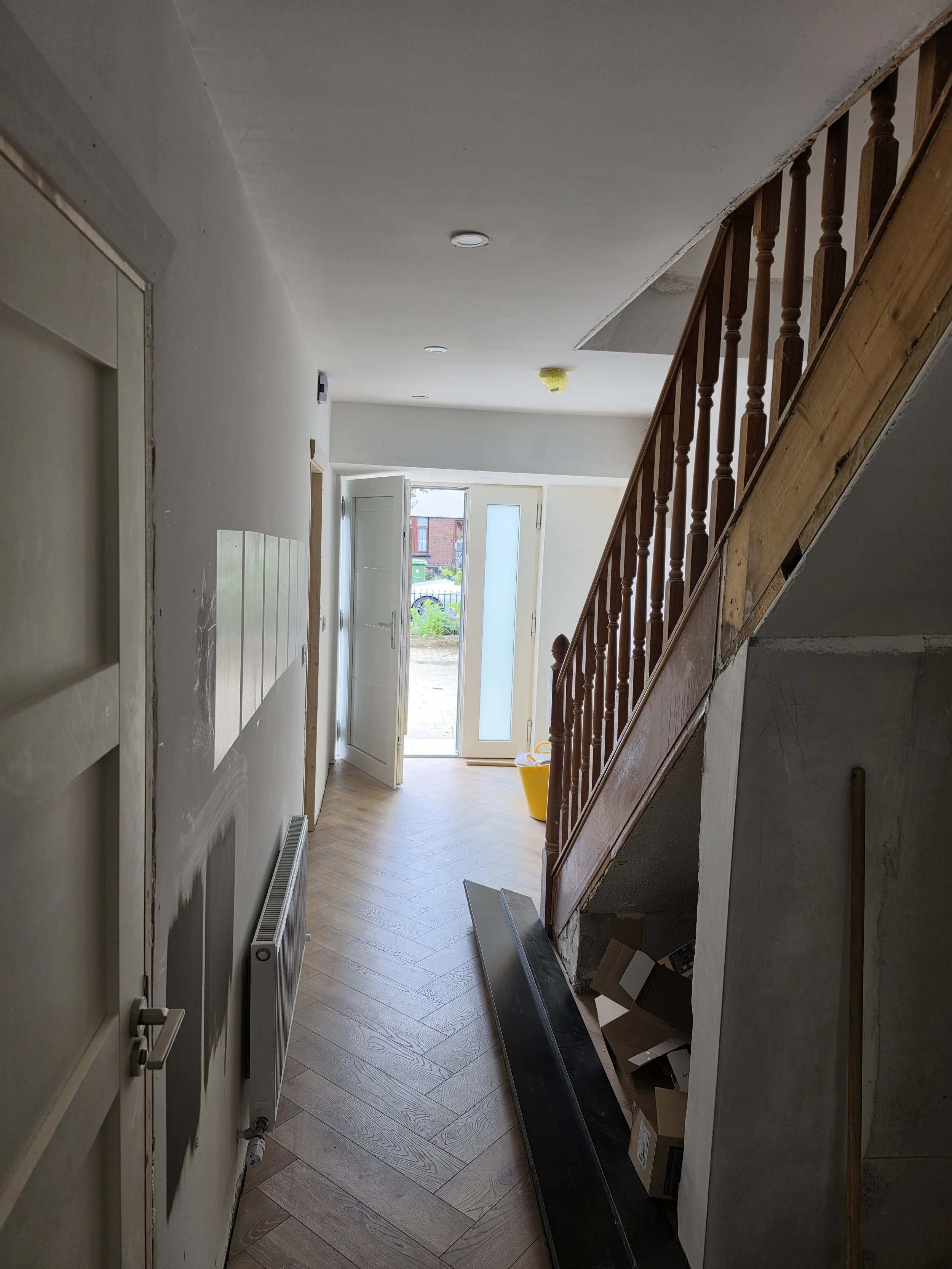 Interior hallway under renovation with a staircase on the right, open door at the end leading outside, and construction materials on the floor.