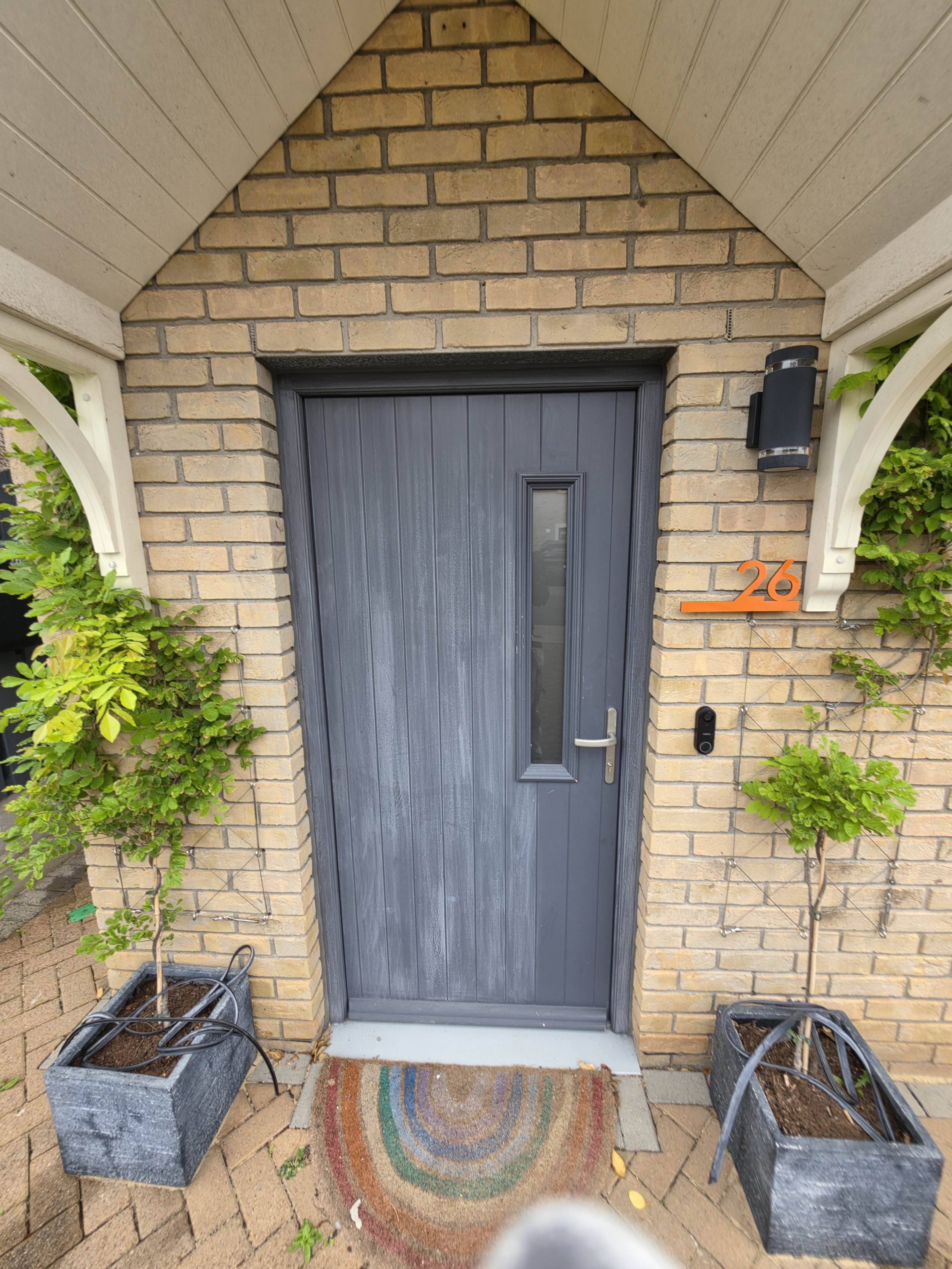 View of a front door with a dark gray wooden door, brick wall, and a colorful semi-circle patterned doormat. Two large planters with soil are placed on either side of the door, each with a small green plant. The house number 26 is displayed in orange on the right side of the wall. There is a black outdoor light fixture and a doorbell or intercom system beside the door.