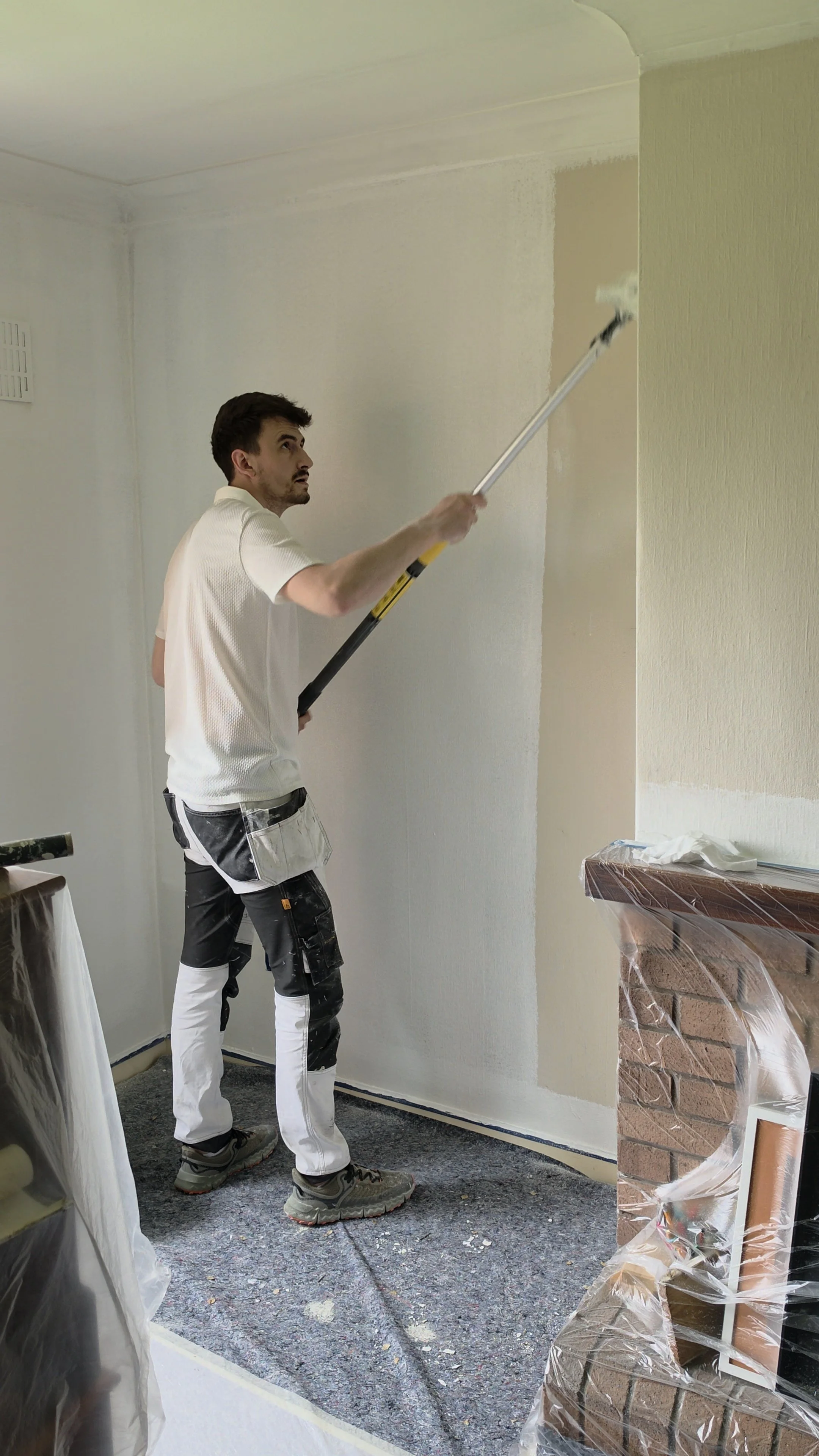 A man painting a wall with a roller brush in a room under renovation; he is wearing paint-splattered work clothes and a tool belt, with protective coverings visible on furniture and fireplace.