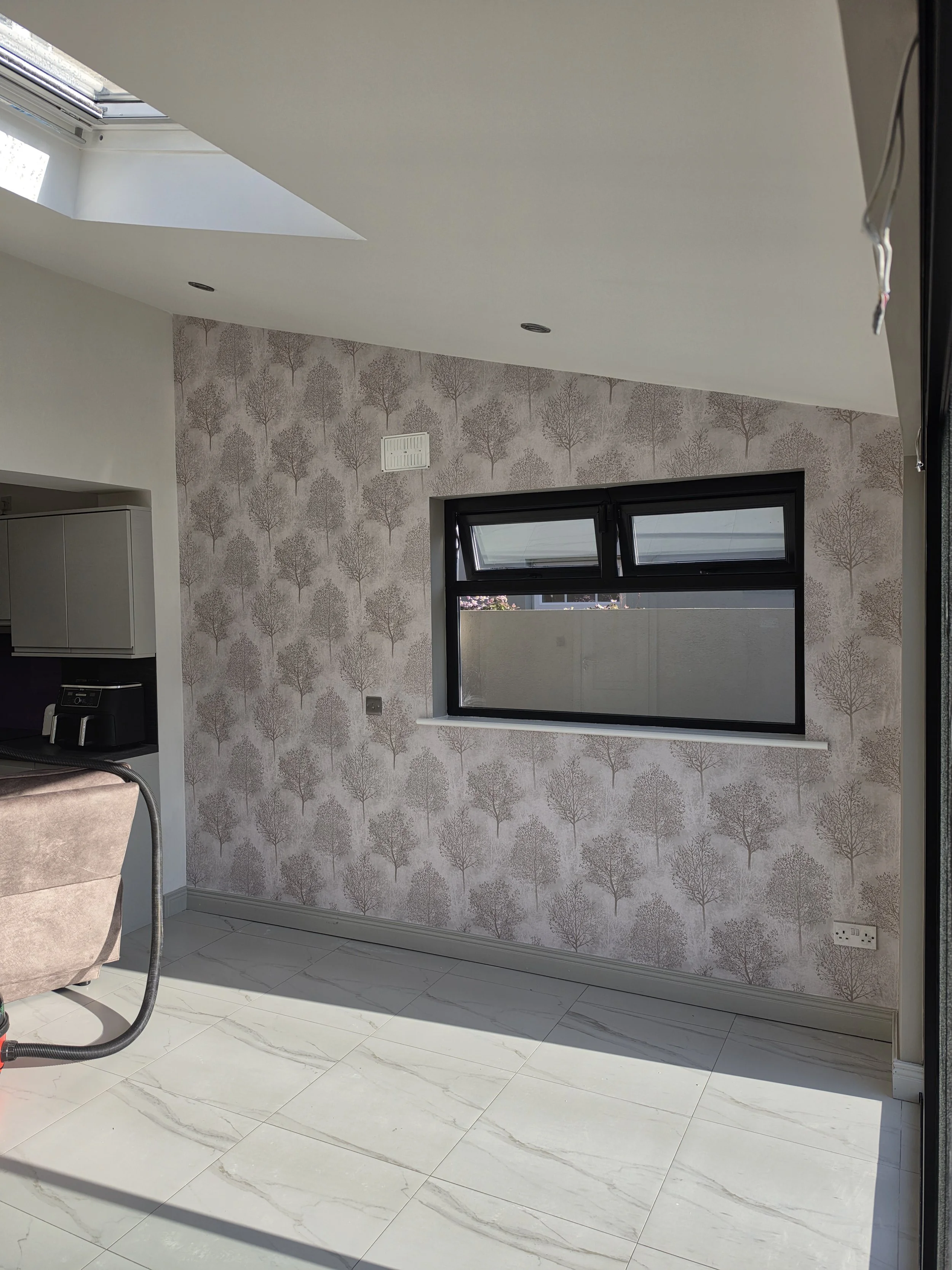 Interior room with a window, patterned wallpaper with trees, and a white tiled floor. Part of a beige armchair and a vacuum cleaner are visible.