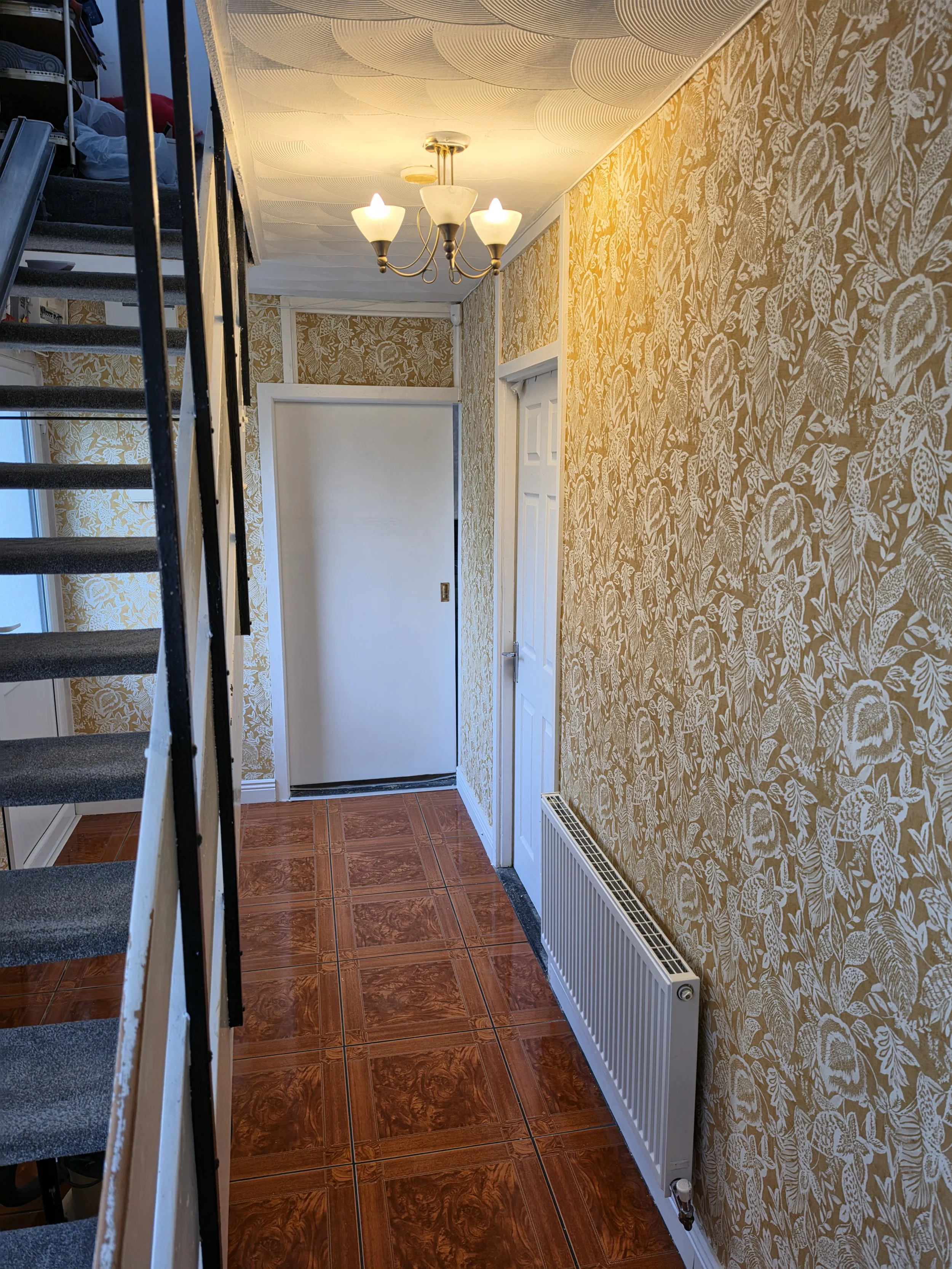 Interior view of a narrow hallway with floral wallpaper on the walls, a white door, a decorative chandelier with three lights, a radiator, tiled wooden floor, and a staircase on the left side.