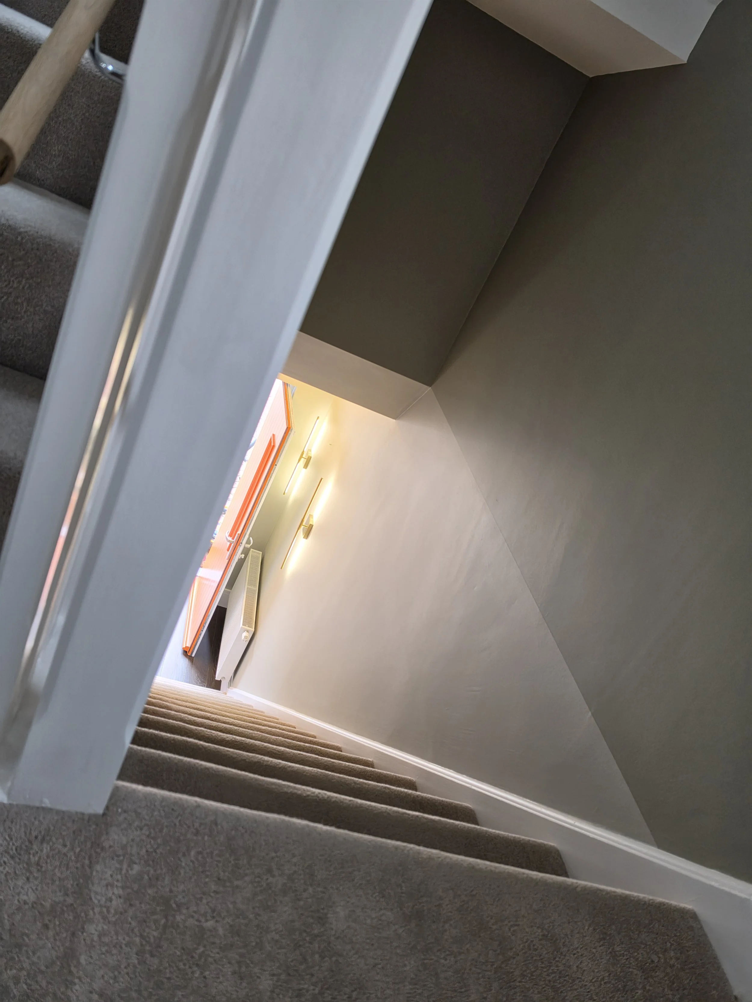 A staircase with carpeted steps viewed from above, leading down to a hallway with modern wall lighting and a door with orange trim.