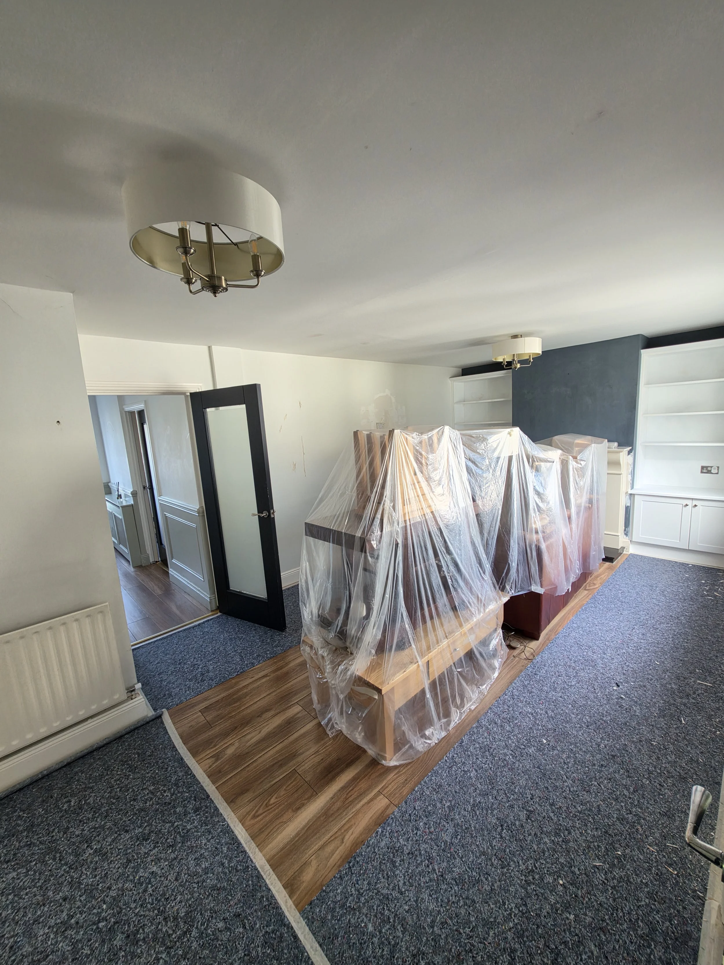 Living room with a removed fireplace, covered furniture, and construction plastic, featuring a mixed carpet and wooden flooring, with white and blue walls and a ceiling-mounted light fixture.