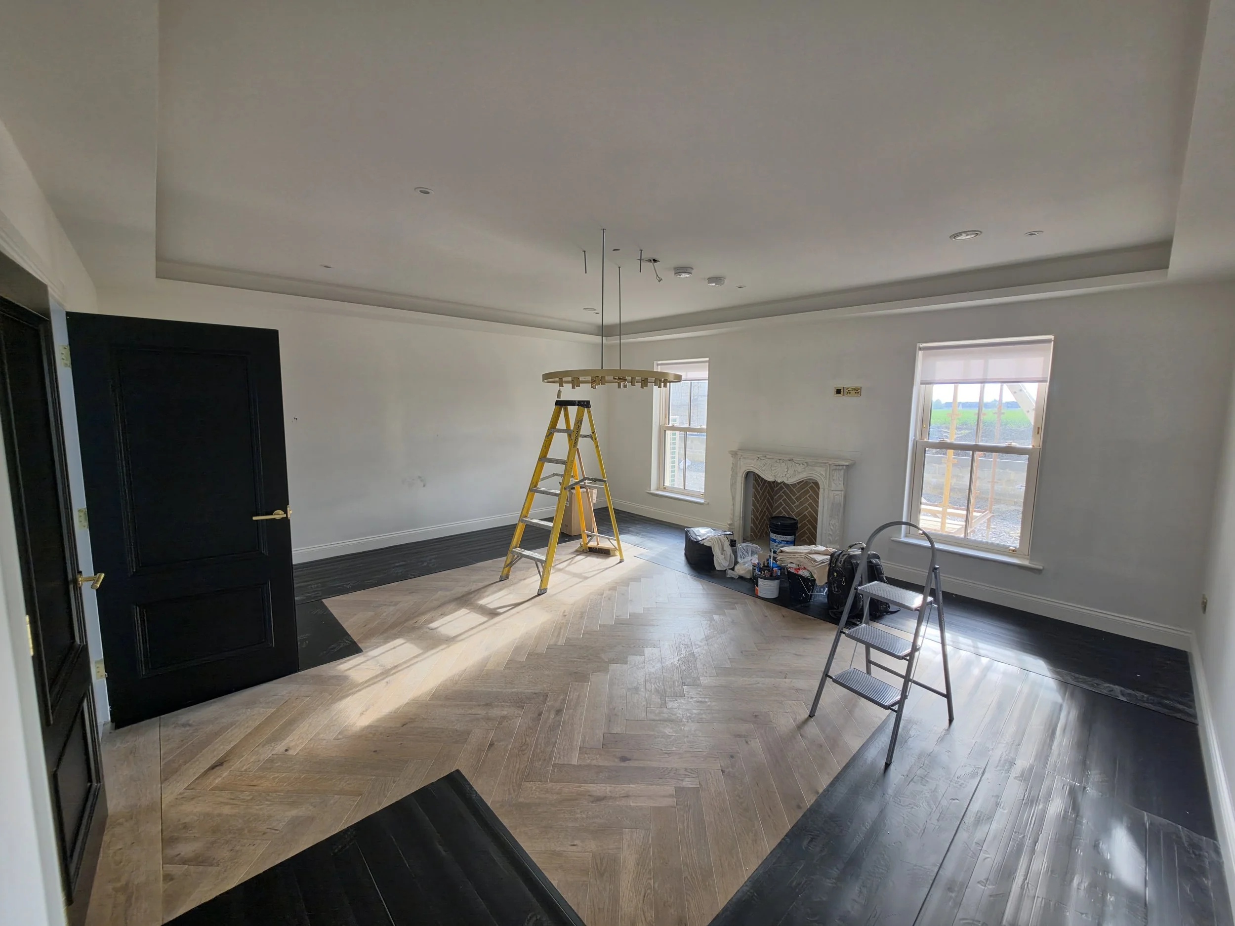 Living room with a ceiling that is being renovated, containing a ladder, tools, and construction supplies, with hardwood floors, windows, and a fireplace.