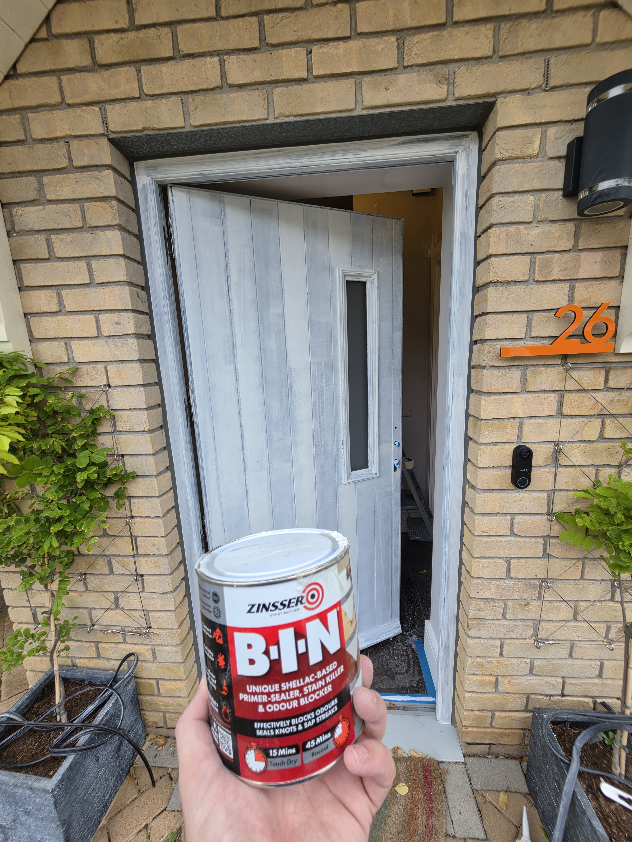 A person holding a can of Zinsser BIN shellac-based primer/dealer/stain blocker in front of a house entrance with a brick wall, doorway, and a house number 26 on an orange sign.