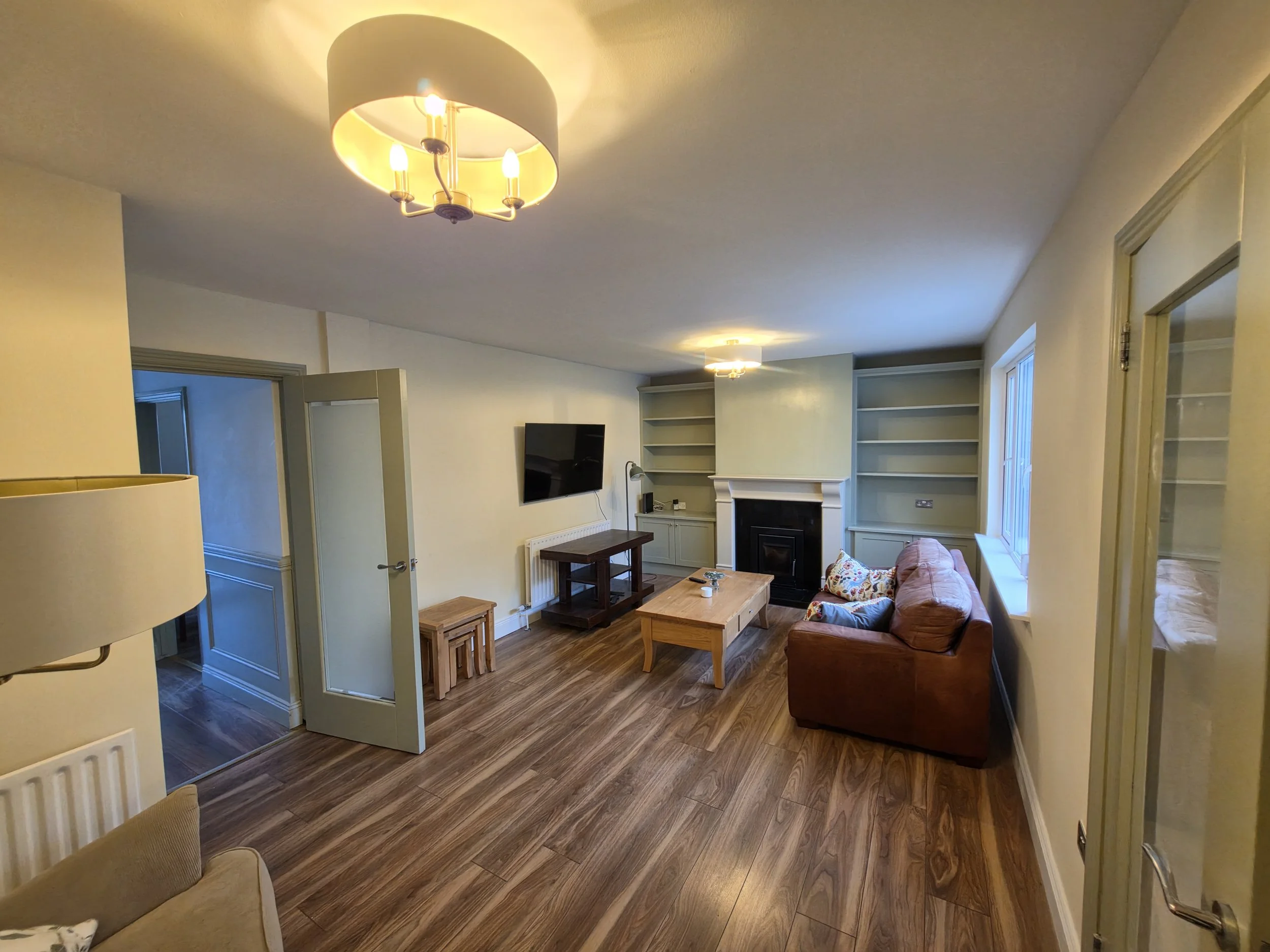 Living room with a brown sofa, a wooden coffee table, a wall-mounted TV, built-in bookshelves, and ceiling lights.