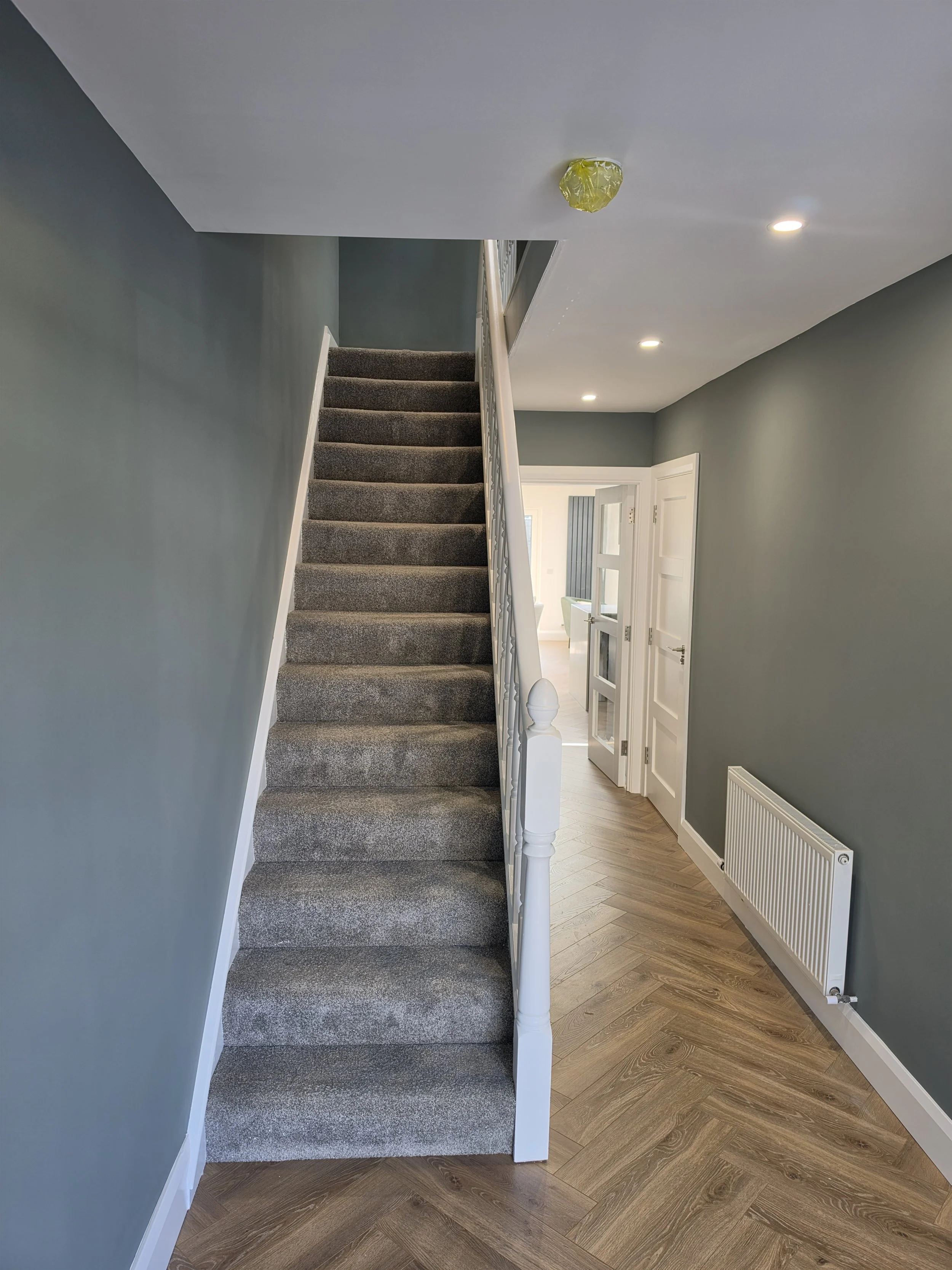 Image of a staircase with carpeted steps and a white handrail on the right side, leading to the upper floor. The walls are painted a muted blue-gray, with a section showing a white door with glass panes on the right side. Recessed ceiling lights illuminate the space, and a radiator is installed on the wall near the door. The flooring near the staircase appears to be wooden in a herringbone pattern.