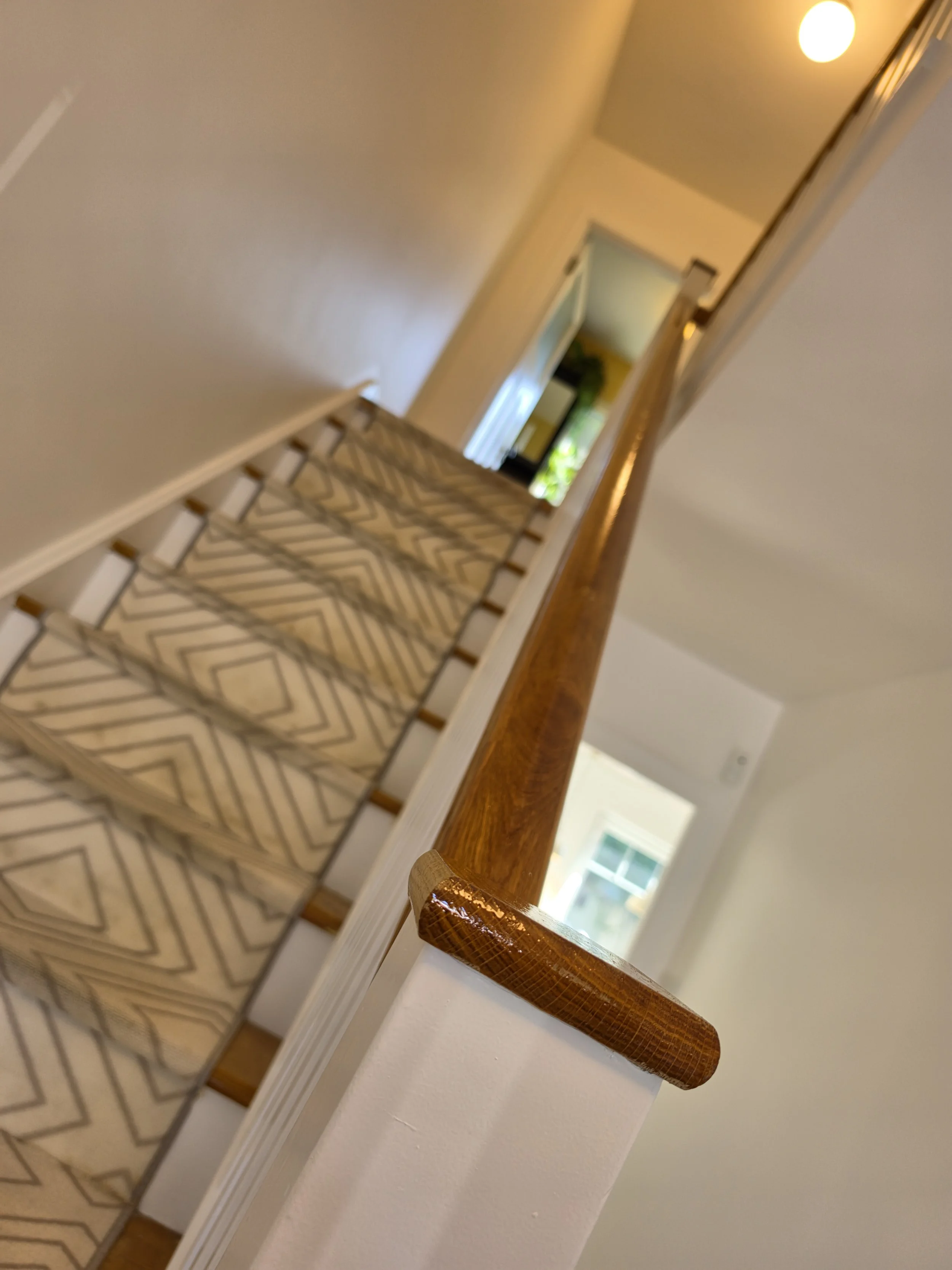 View down a staircase with a patterned carpet runner, a wooden handrail, and a hallway with doorways and a window in the background.