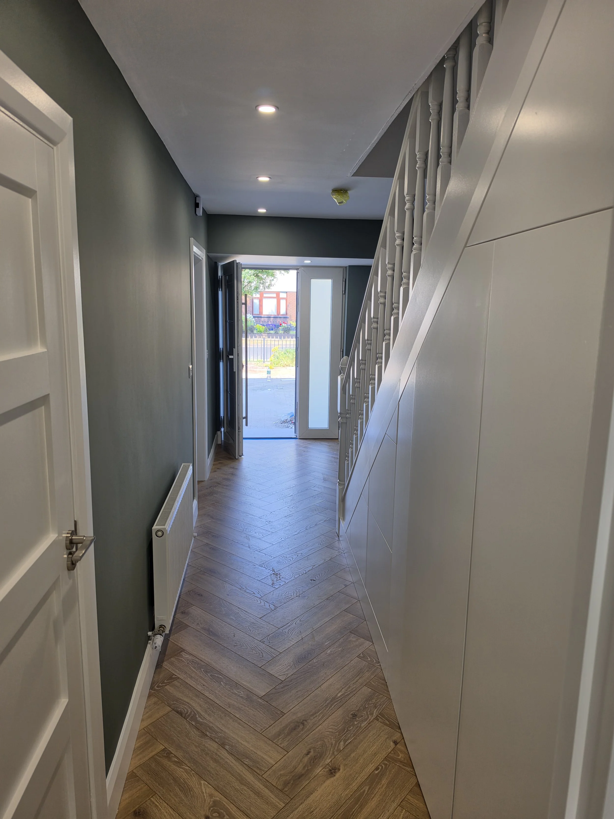 Hallway with wooden floor, green wall on the left, white wall on the right, with stair railing visible, and open front door showing a yard outside.