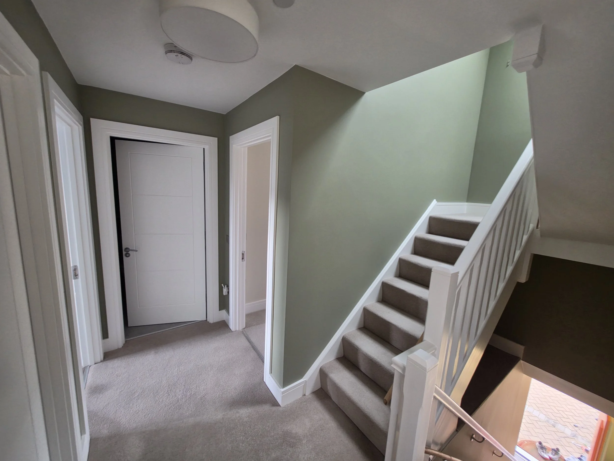 The interior of a house showing a staircase with beige carpet, a white railing, and a light green wall. There are two closed white doors and some hallway space. Part of a kitchen and an outdoor area are visible.