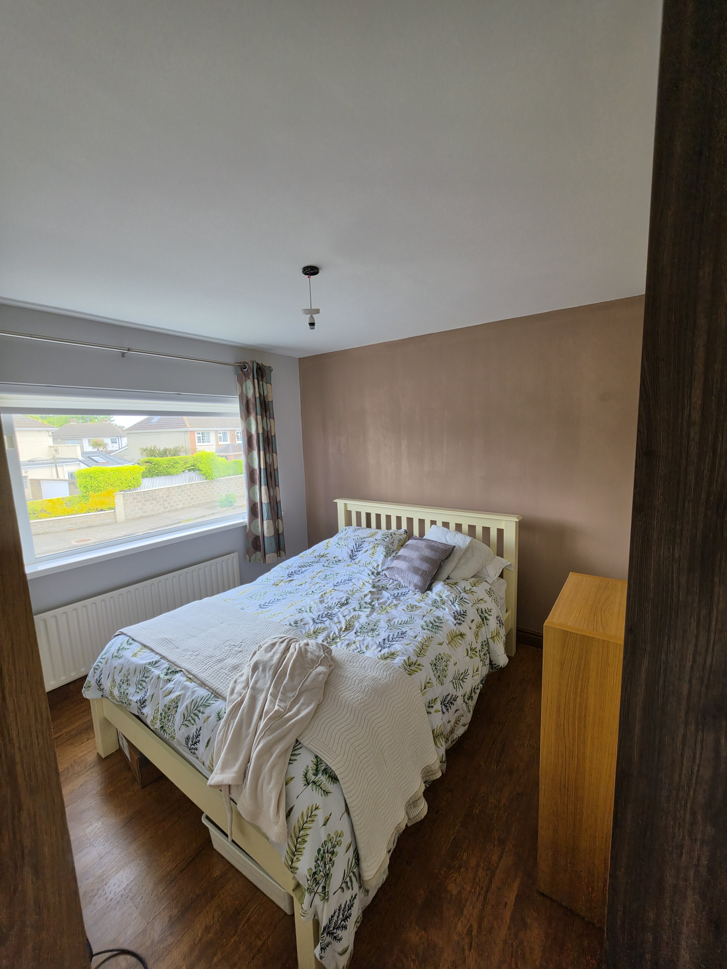 A bedroom with a double bed, white wooden frame, floral bedspread, pillows, a window with polka dot curtains, a small yellow side table, and a ceiling light fixture without a bulb.