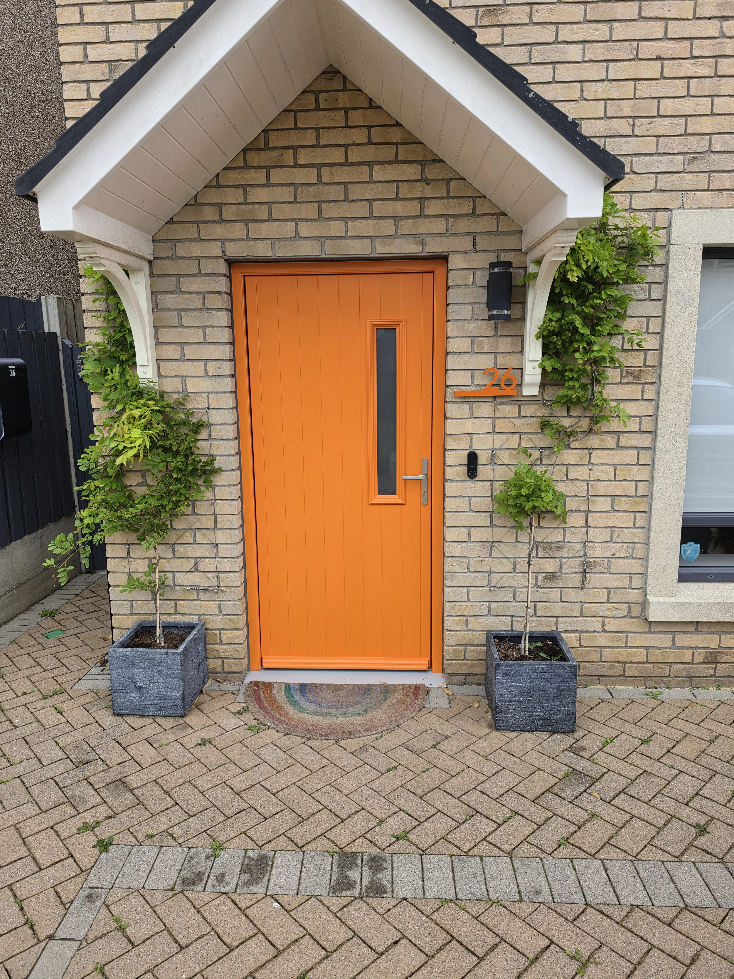 A brick house entrance with an orange front door, black house numbers 26, and two potted green plants. The entry has a small greeting rug, and there's a black mailbox to the left side of the door.