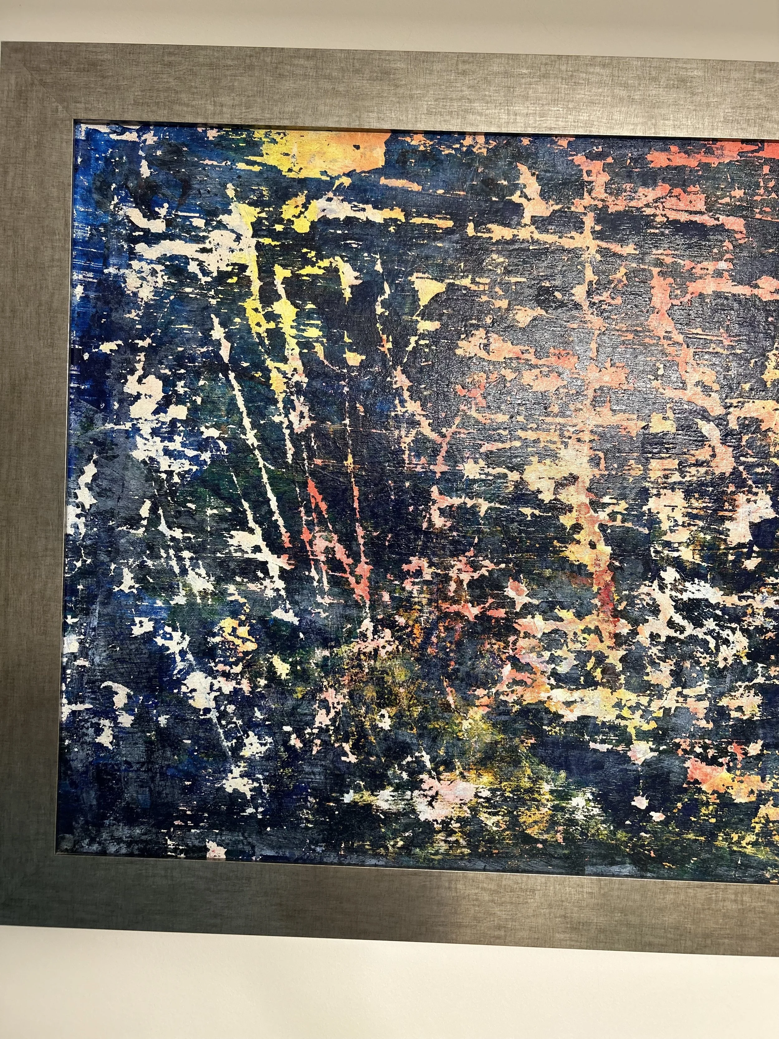 Abstract painting with dark blue, black, yellow, pink, green, and white splashes and streaks, framed and displayed on a wall.