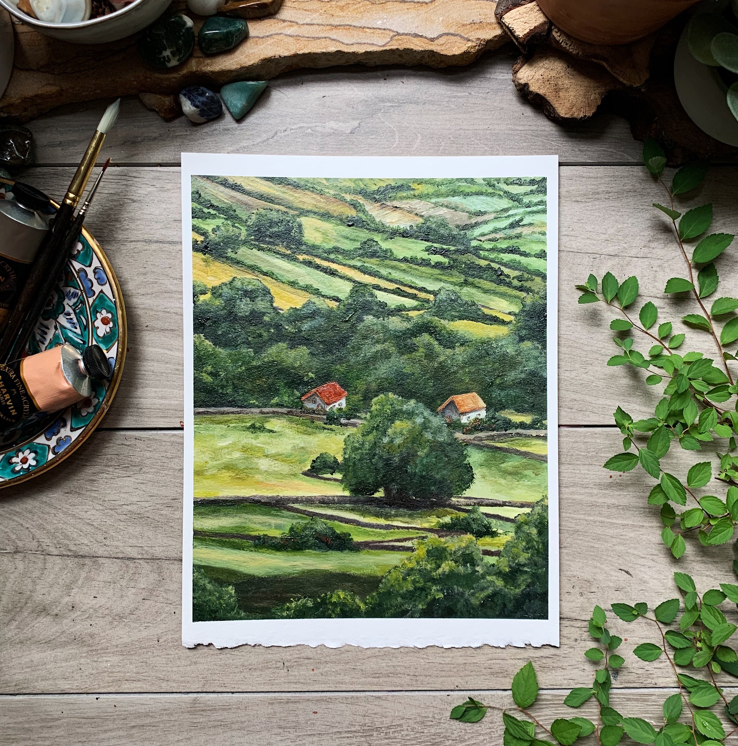 A landscape illustration of rolling green hills with trees and small houses, placed on a wooden surface with decorative stones, a ceramic plate, paintbrushes, and leafy branches surrounding it.