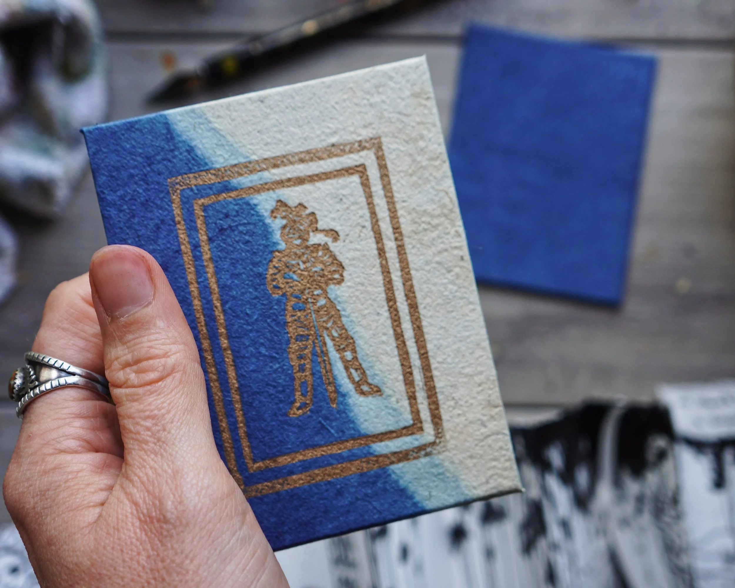A hand holds a small watercolor painting with a blue background, brown border, and a gold outline of a tree, with a blurred background.