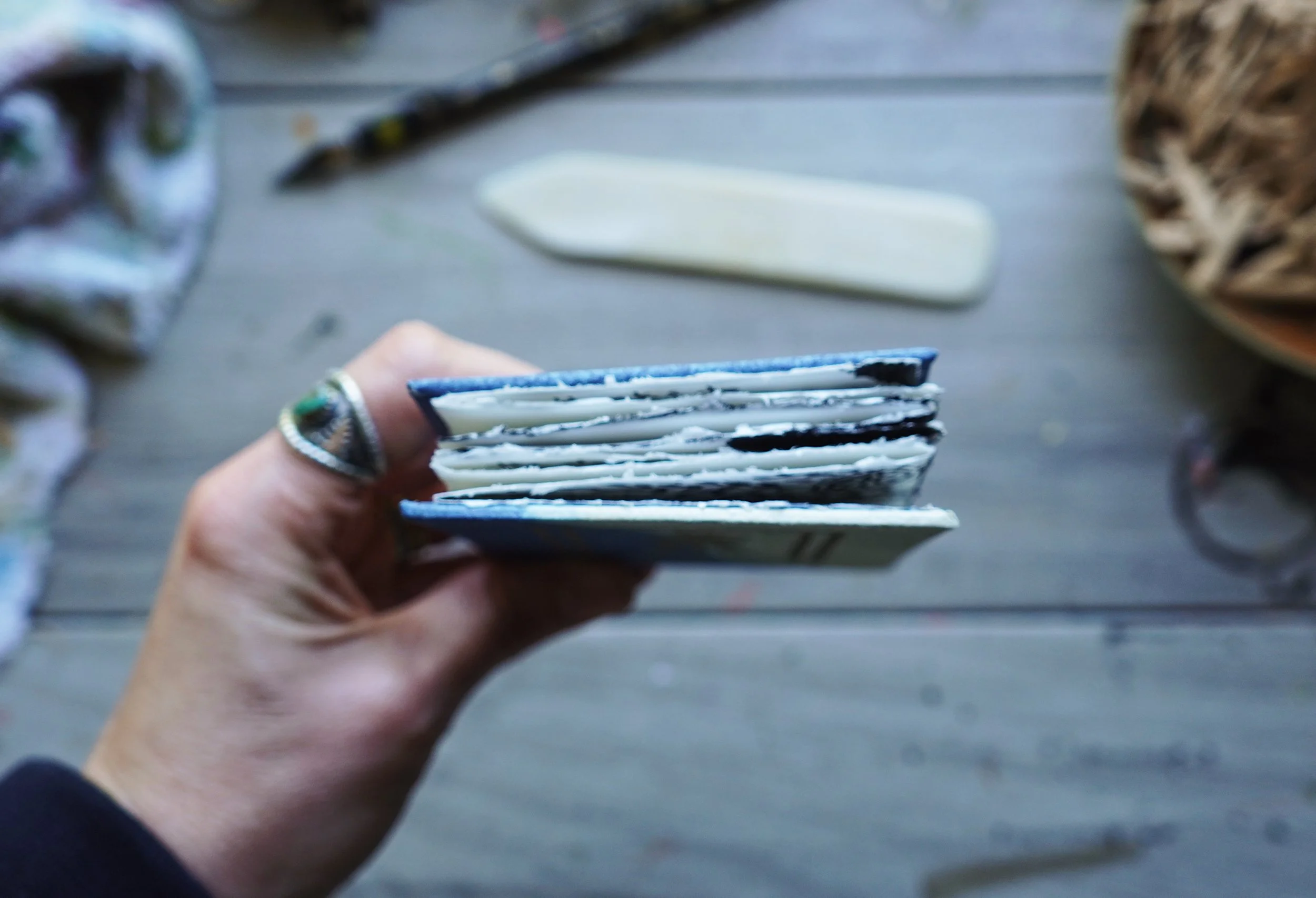 Person holding a small stack of four books with a ring on their finger, with a blurred background of a desk with various objects including a white object and a pencil.