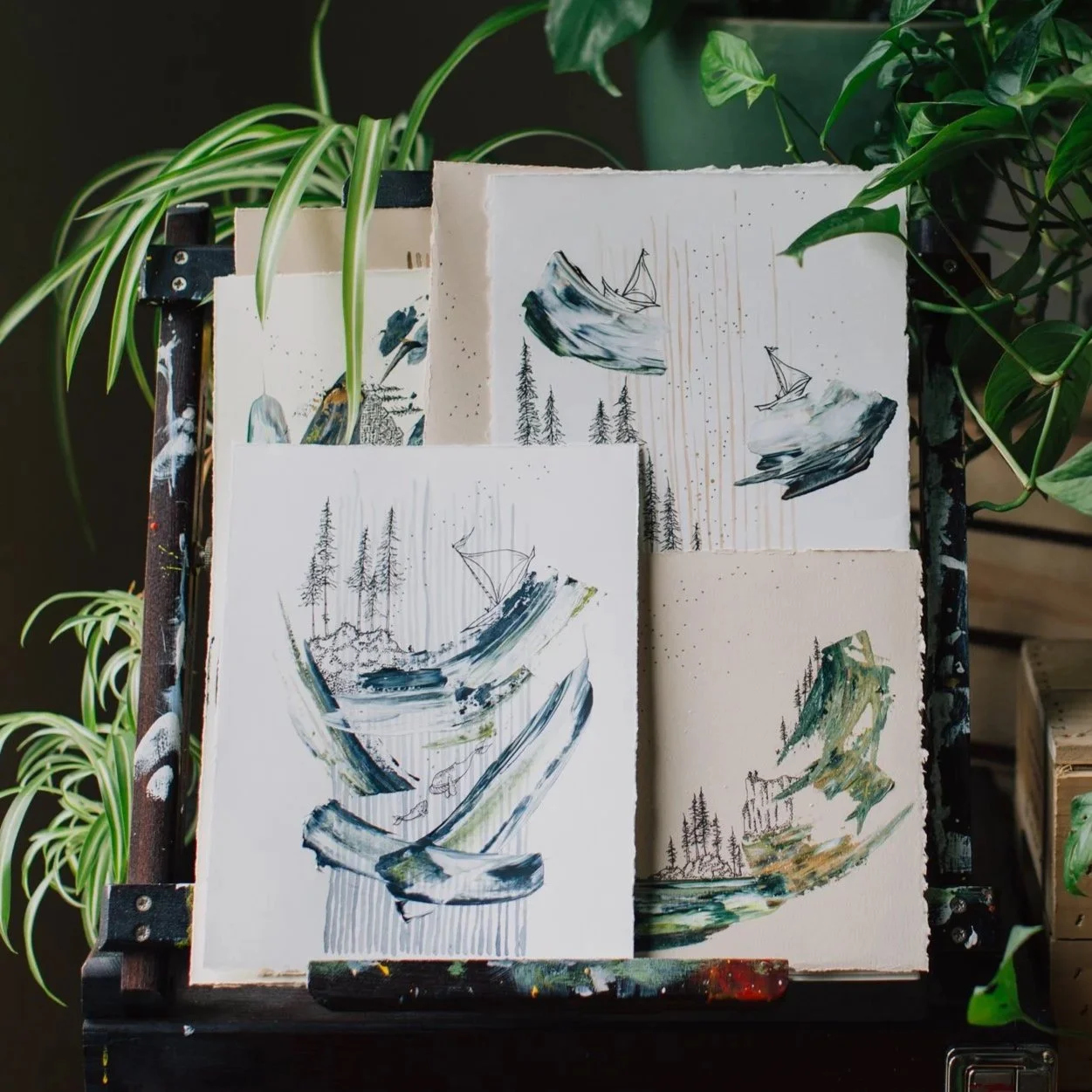 Several small abstract landscape paintings on textured paper, featuring trees and mountains, displayed on a black, paint-splattered easel with green indoor plants in the background.