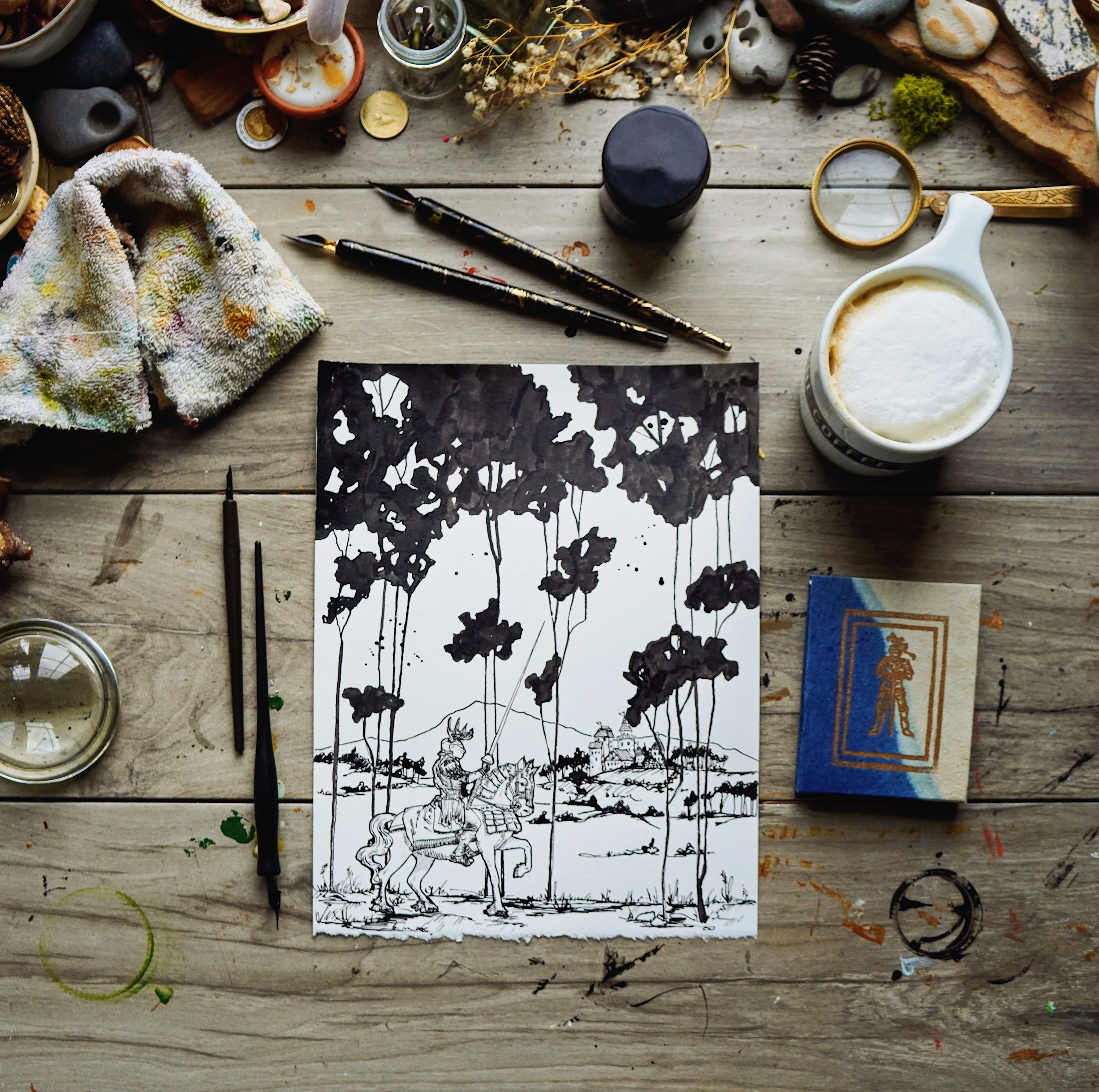 A black ink or paint illustration of a scene with a person on a horse in the foreground, and tall trees, a castle, and a figure in the background, on a piece of paper on a wooden table surrounded by various art supplies and objects.