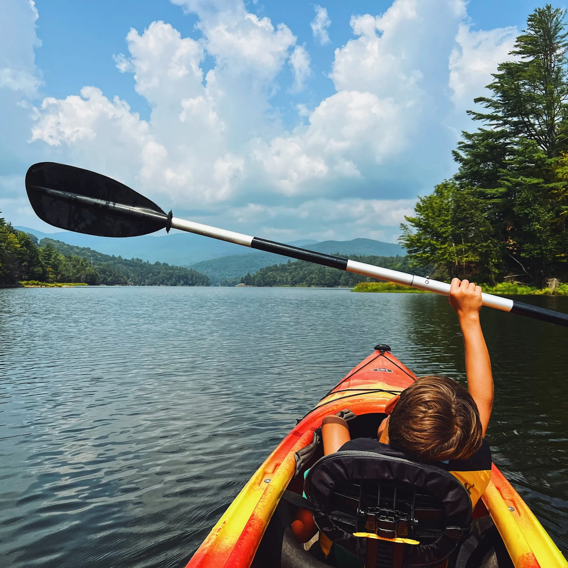 Child kayaking on a lake surrounded by trees and mountains under a partly cloudy sky, with a paddle raised in the air.