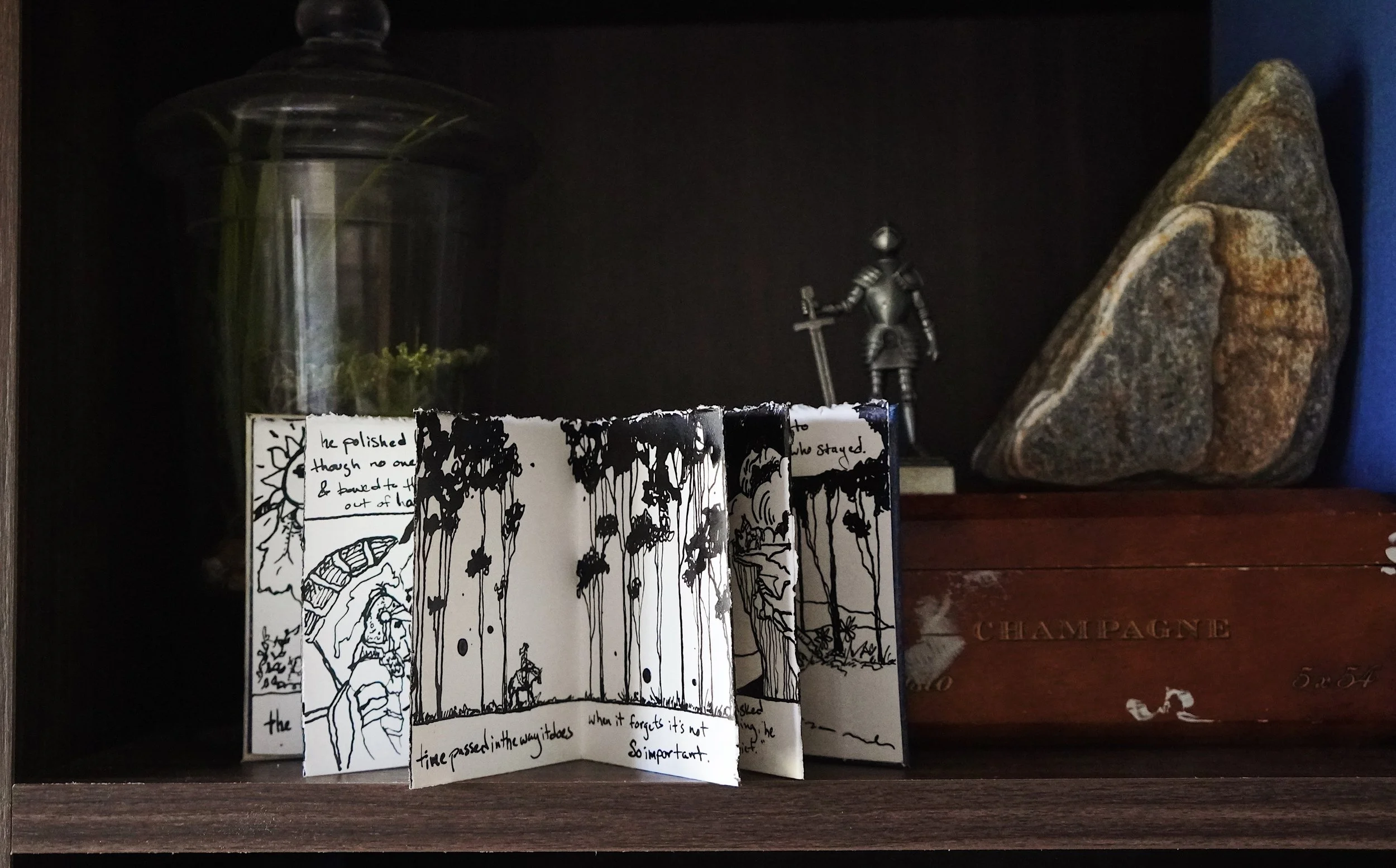 A black and white illustrated folded paper booklet with hand-written text and drawings, placed on a dark wooden shelf. Behind the booklet, there is a large glass container partially visible on the left, a small metallic figurine of a knight with a sw
