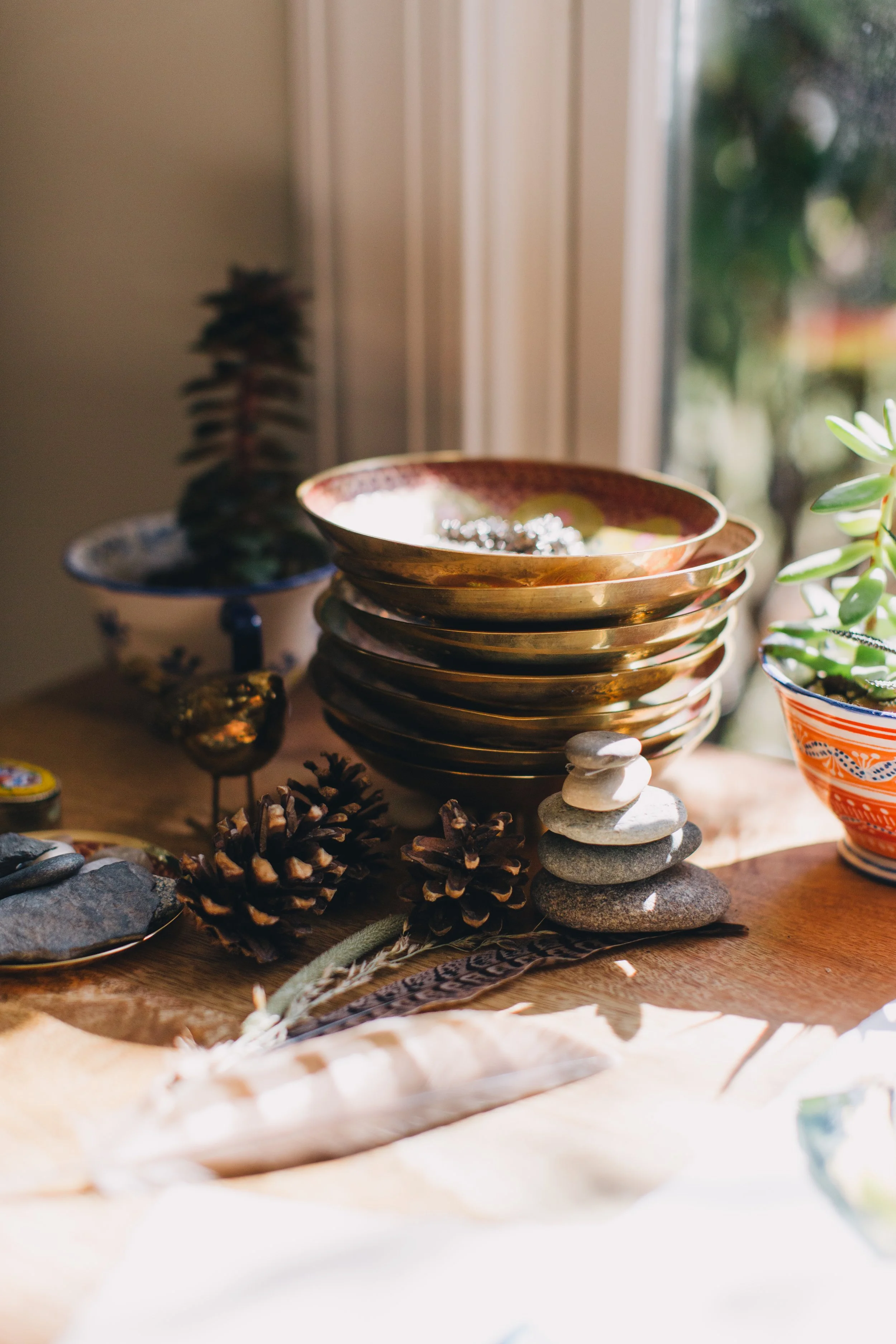 Stack of decorative bowls, pinecones, rocks, and a feather on a wooden surface by a window with natural light.