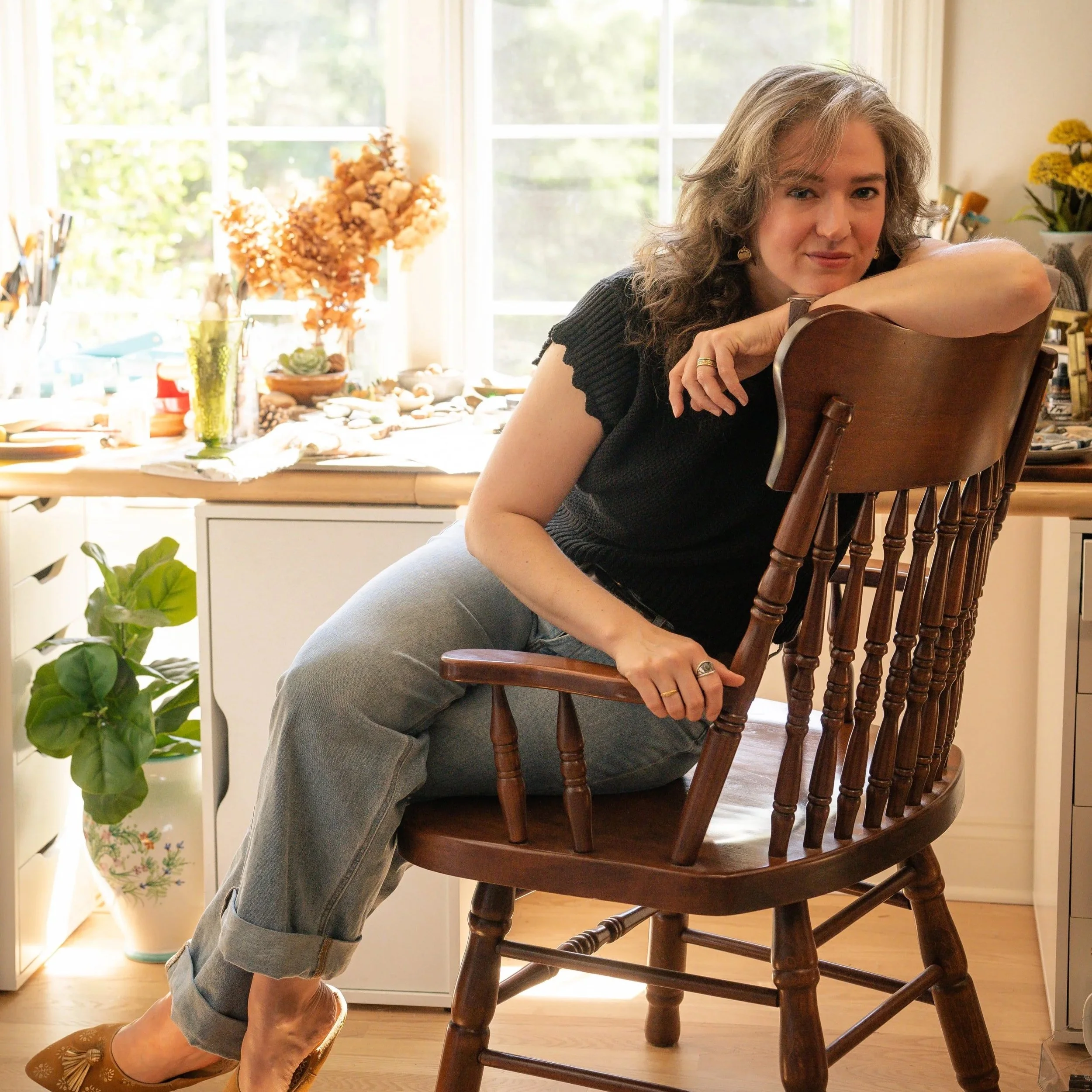 A woman leaning on a wooden chair in her kitchen, with her head resting on her arms. She's wearing a black top and gray pants, with a nose ring and earrings. The background shows a table with decorative stones, a green plant, and a window with sunlight streaming in, illuminating the space.