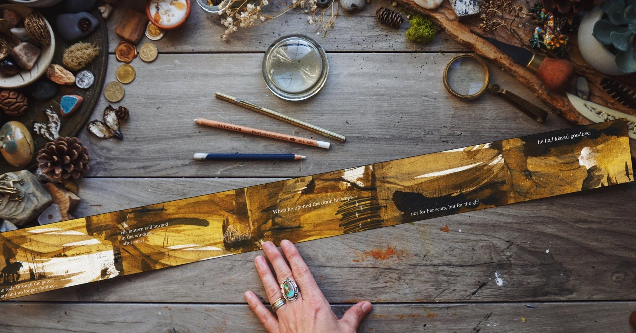 A person's hand with rings holding a printed storybook or illustrated paper on a wooden table surrounded by various small objects, including pencils, a magnifying glass, pinecones, stones, and decorative items, with a part of the story visible in the
