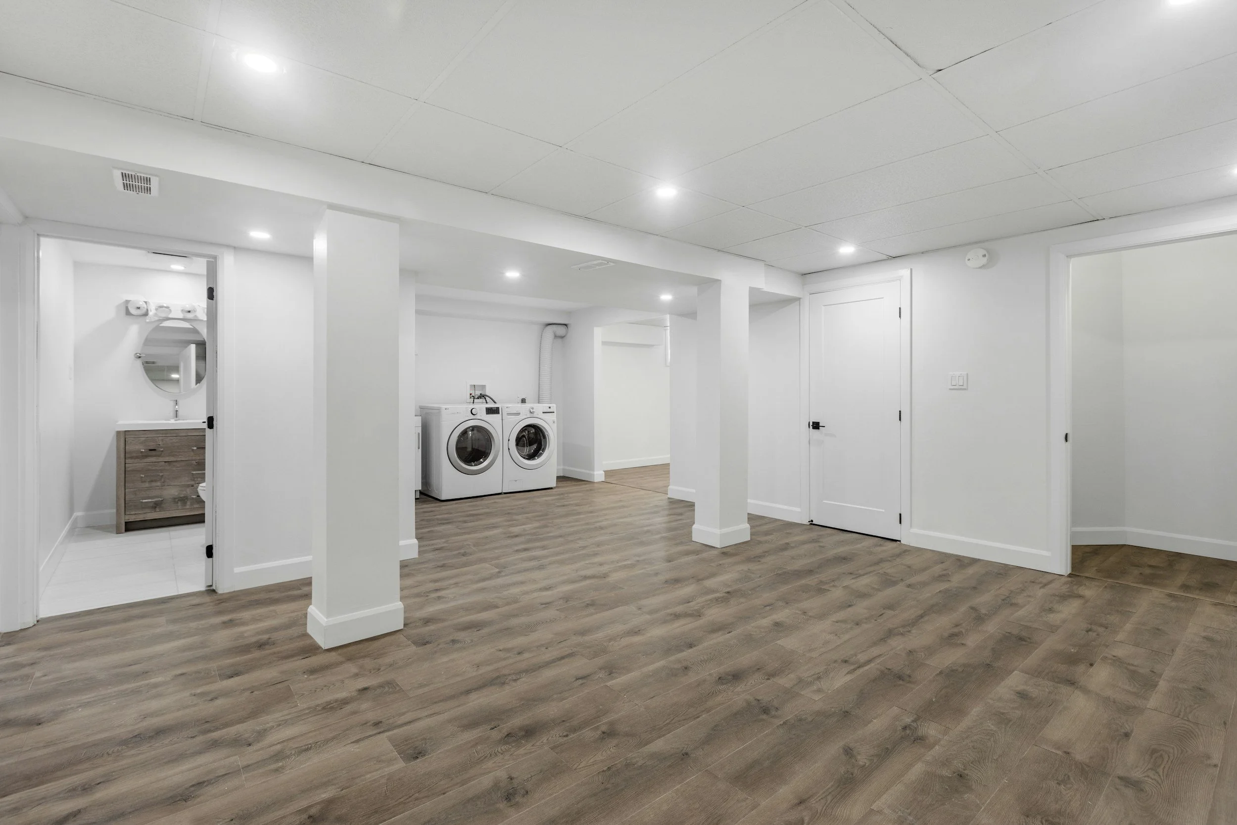 A spacious basement laundry room with white walls, a white ceiling with recessed lighting, a washer and dryer set, wood-like flooring, and a doorway leading to a bathroom with a mirror and wooden cabinet.