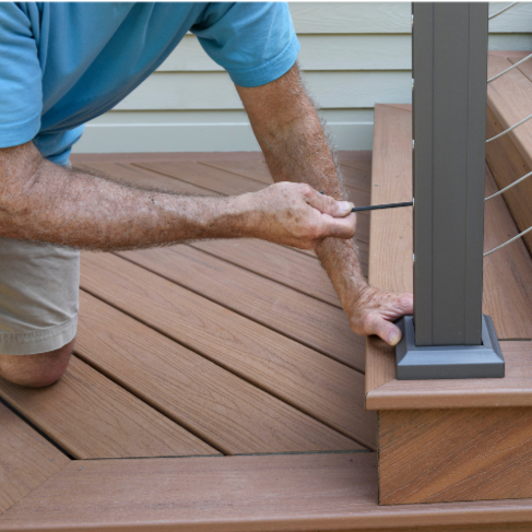 A person kneeling on a wooden deck, tightening a screw on a railing post with a screwdriver.