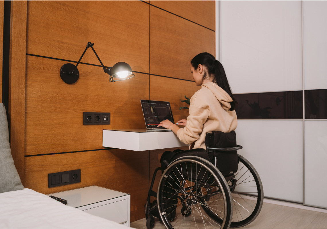 A woman in a wheelchair is working on a laptop at a floating desk in a modern bedroom with wooden panel walls.