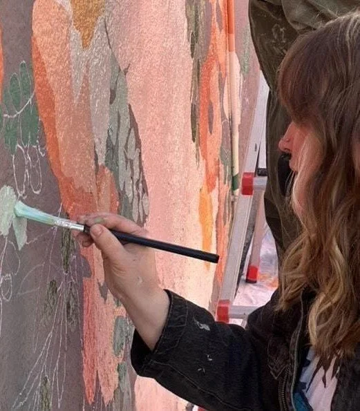 A woman painting on a colorful outdoor mural with a brush.
