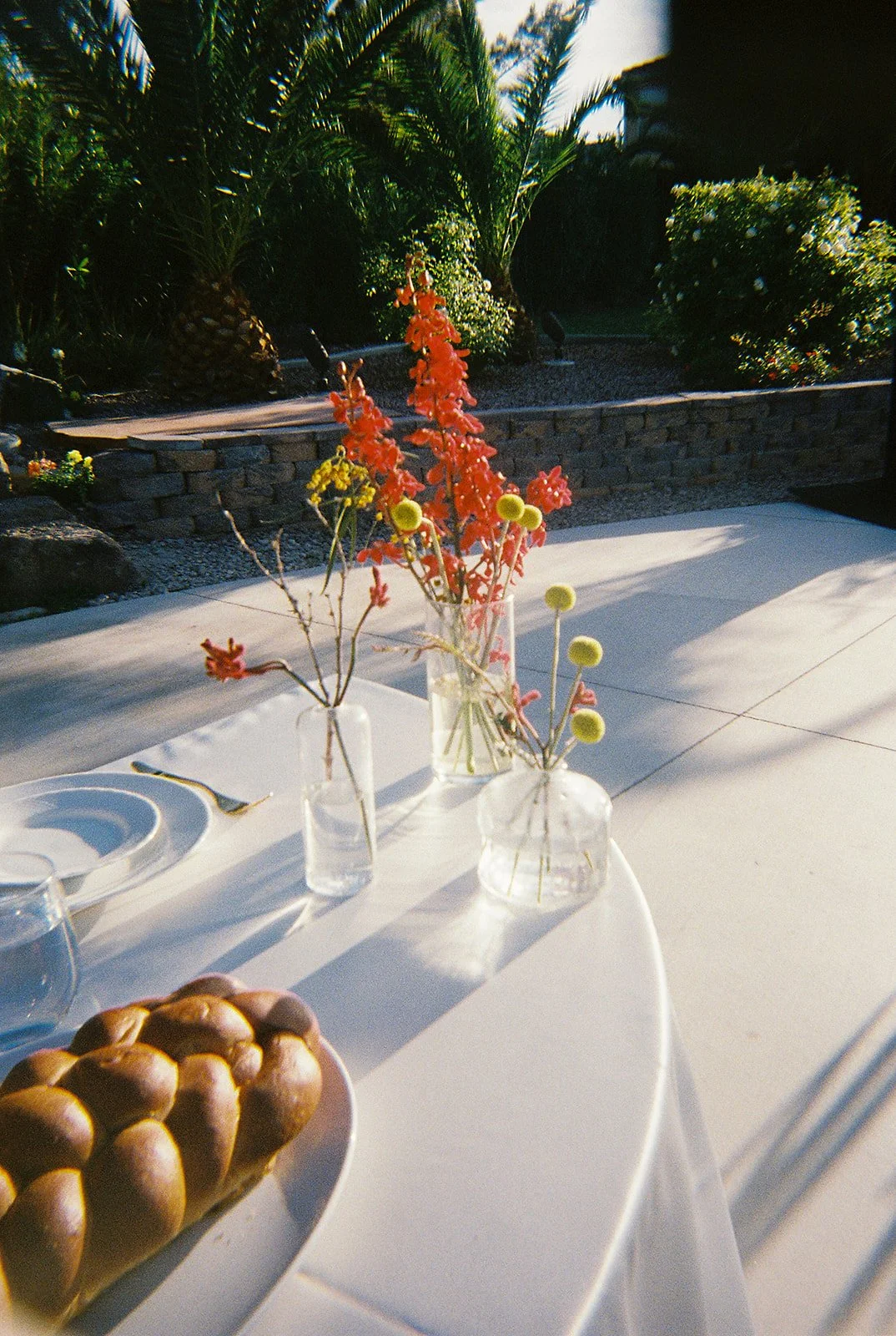 Mesa decorada con floreros de vidrio con flores coloridas y un plato con pan en un entorno exterior, con plantas y árboles de fondo.