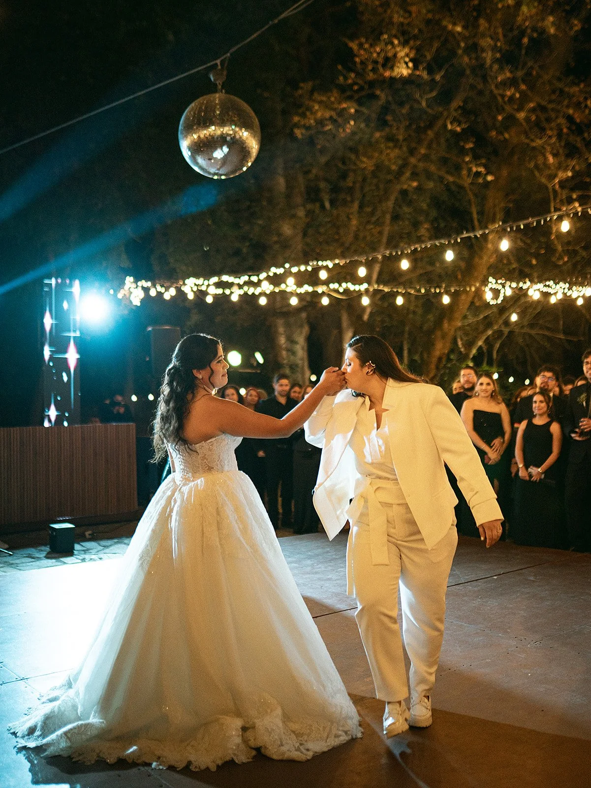 Dos personas bailando en una fiesta al aire libre de noche, una con vestido de novia y la otra con traje, rodeados de invitados, decoraciones de luces y una bola de espejos en el techo.