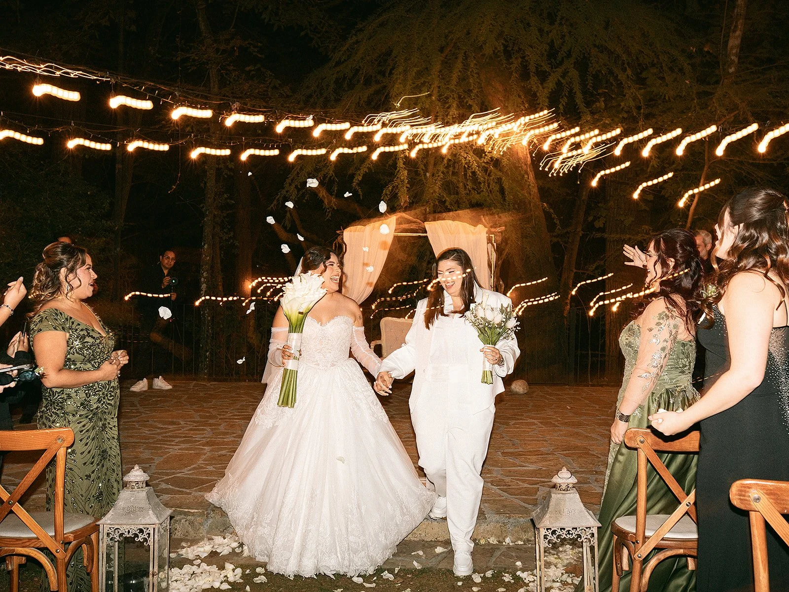 Pareja de novias en una celebración al aire libre, con invitados a su alrededor y decoraciones con luces. Una novia con vestido de boda y otra con ropa blanca, ambas sosteniendo ramos de flores, son las protagonistas del momento.