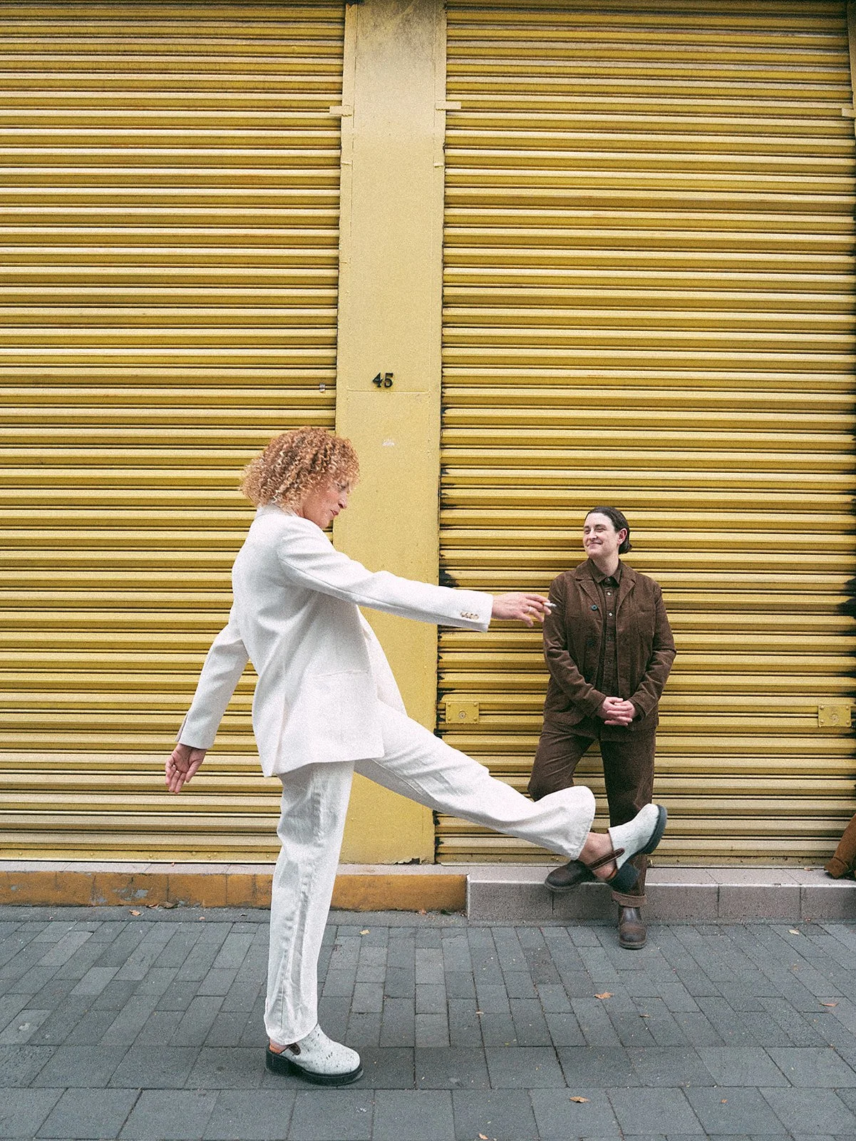 Dos mujeres en una calle, una de ellas de espaldas, usando ropa blanca y la otra de frente, usando ropa marrón, saludándose frente a un fondo de puertas de metal amarillas.