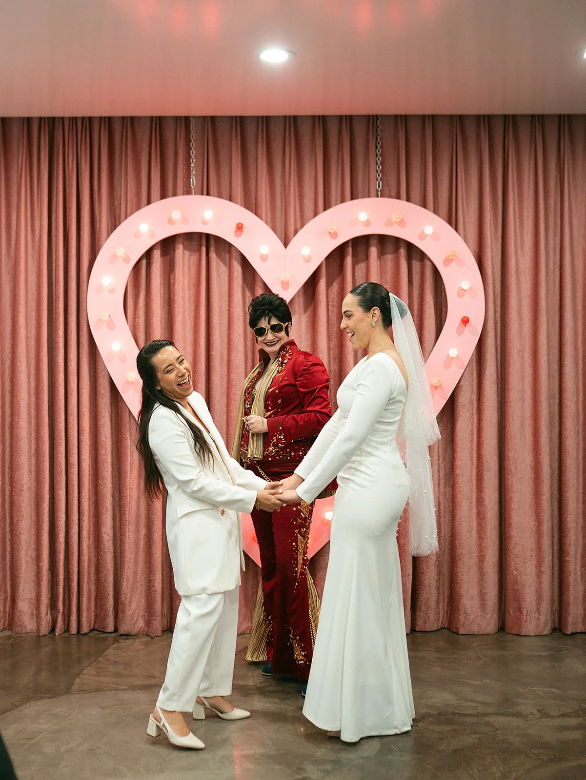 Tres mujeres en una boda, una con vestido blanco de novia y otra con traje blanco, sosteniéndose de las manos y sonriendo, una mujer en el medio con ropa roja y gafas oscuras, con un fondo de cortinas rosadas y un corazón de luces